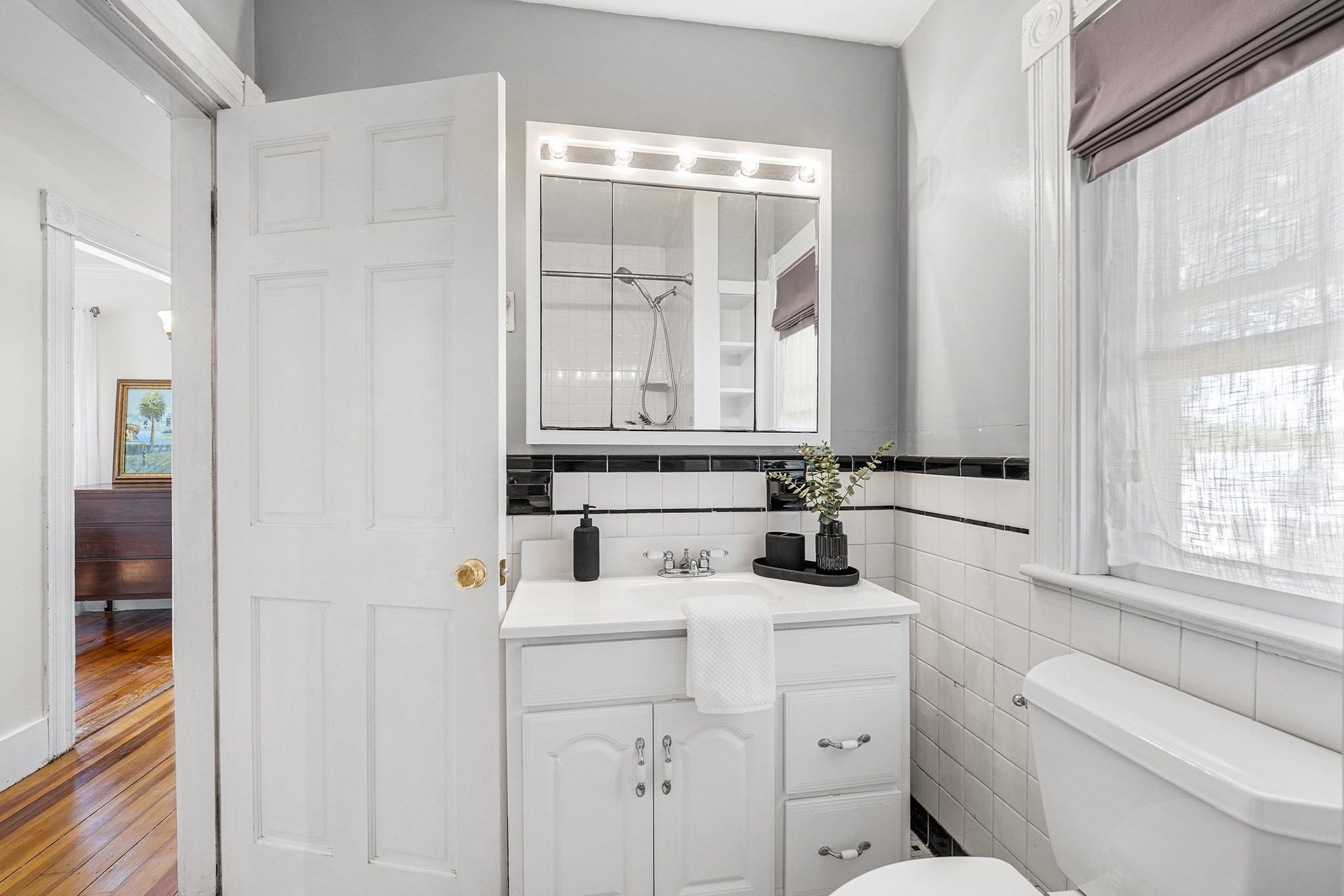 Bathroom with a white vanity, lighted mirrored cabinet, black tile border, and bathtub beside a window.