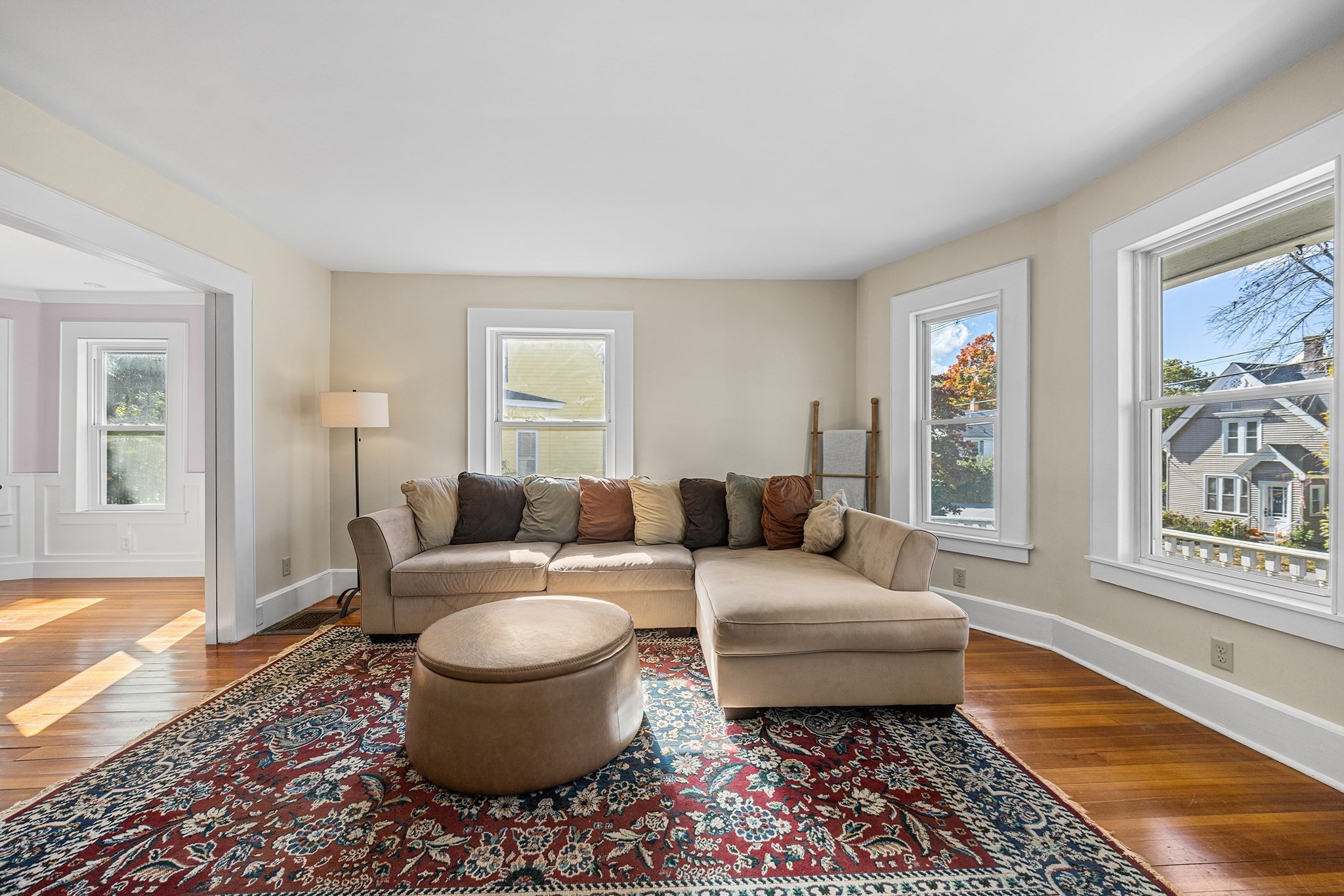 Living room with a beige sectional, patterned red rug, hardwood floors, and bright windows.