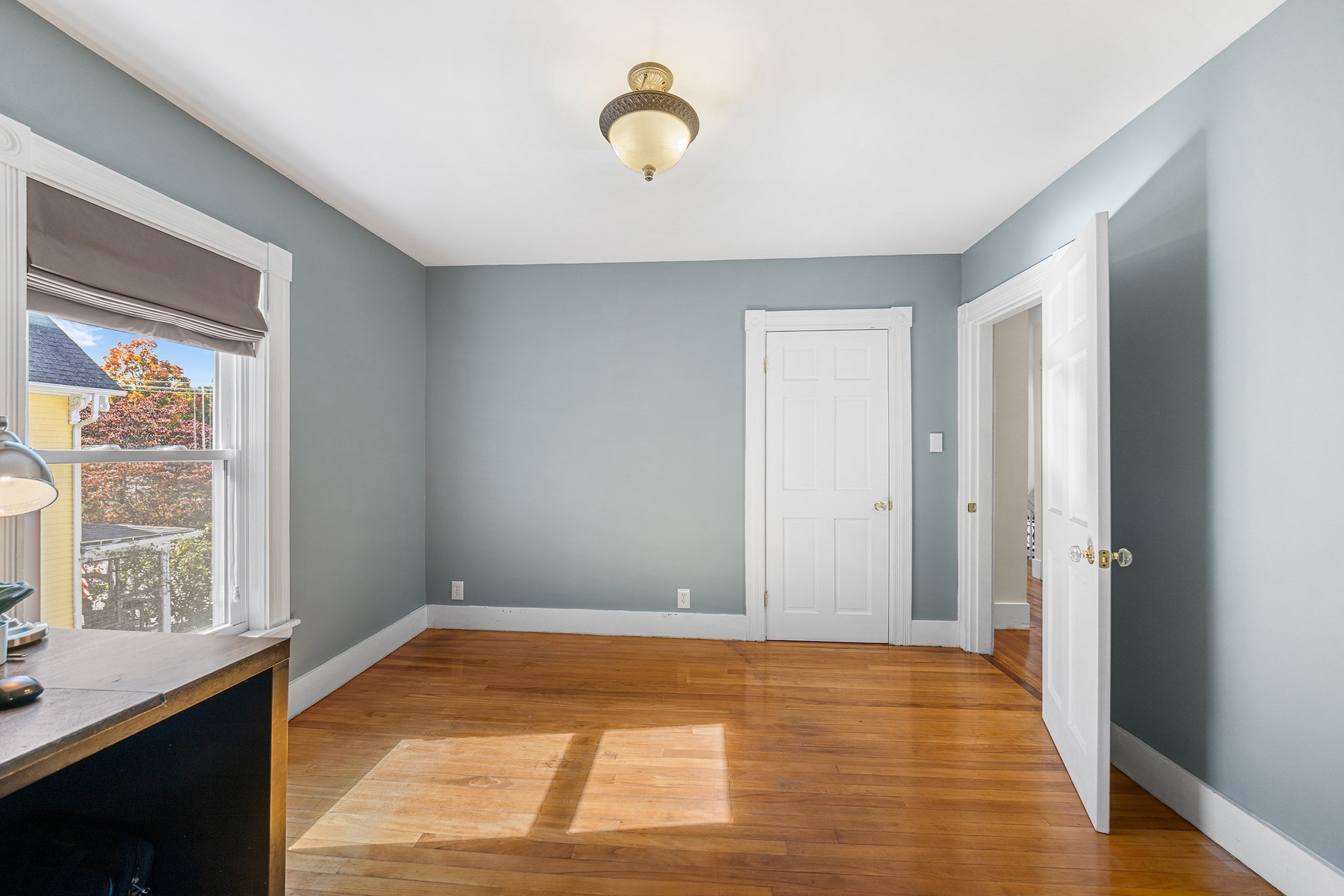 Mostly empty blue-gray room with hardwood floors, white-trimmed doors, and sunlight through a shaded window.