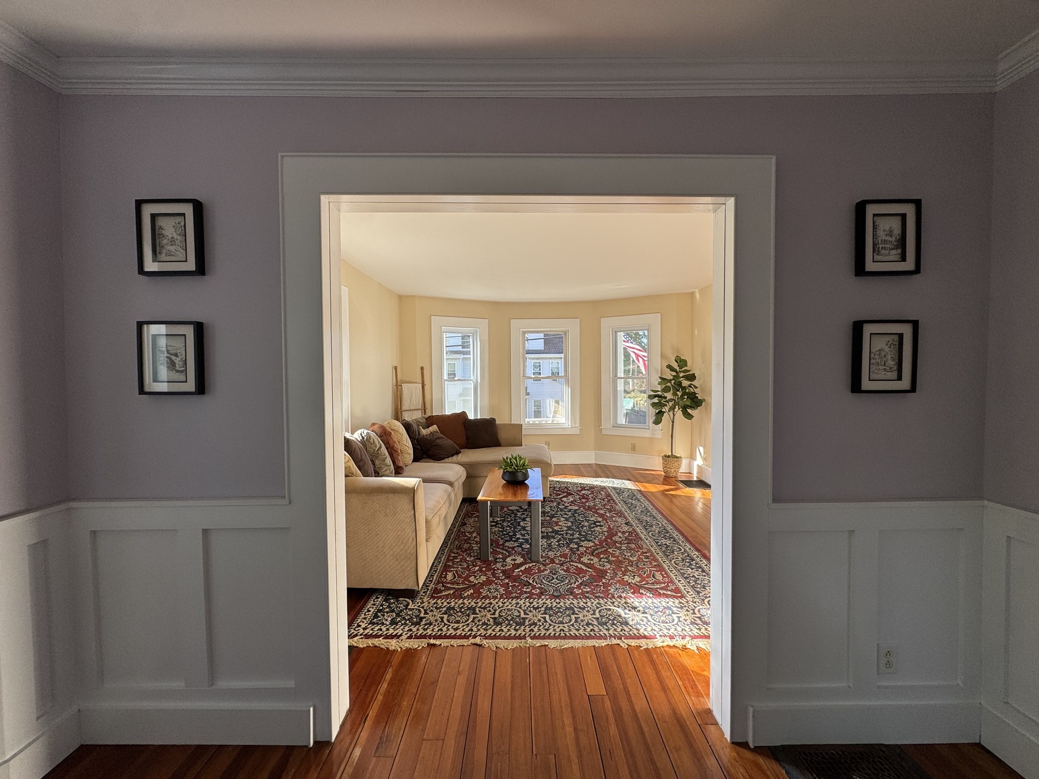 Living room with a beige sectional and patterned rug, seen through a paneled doorway toward three windows.