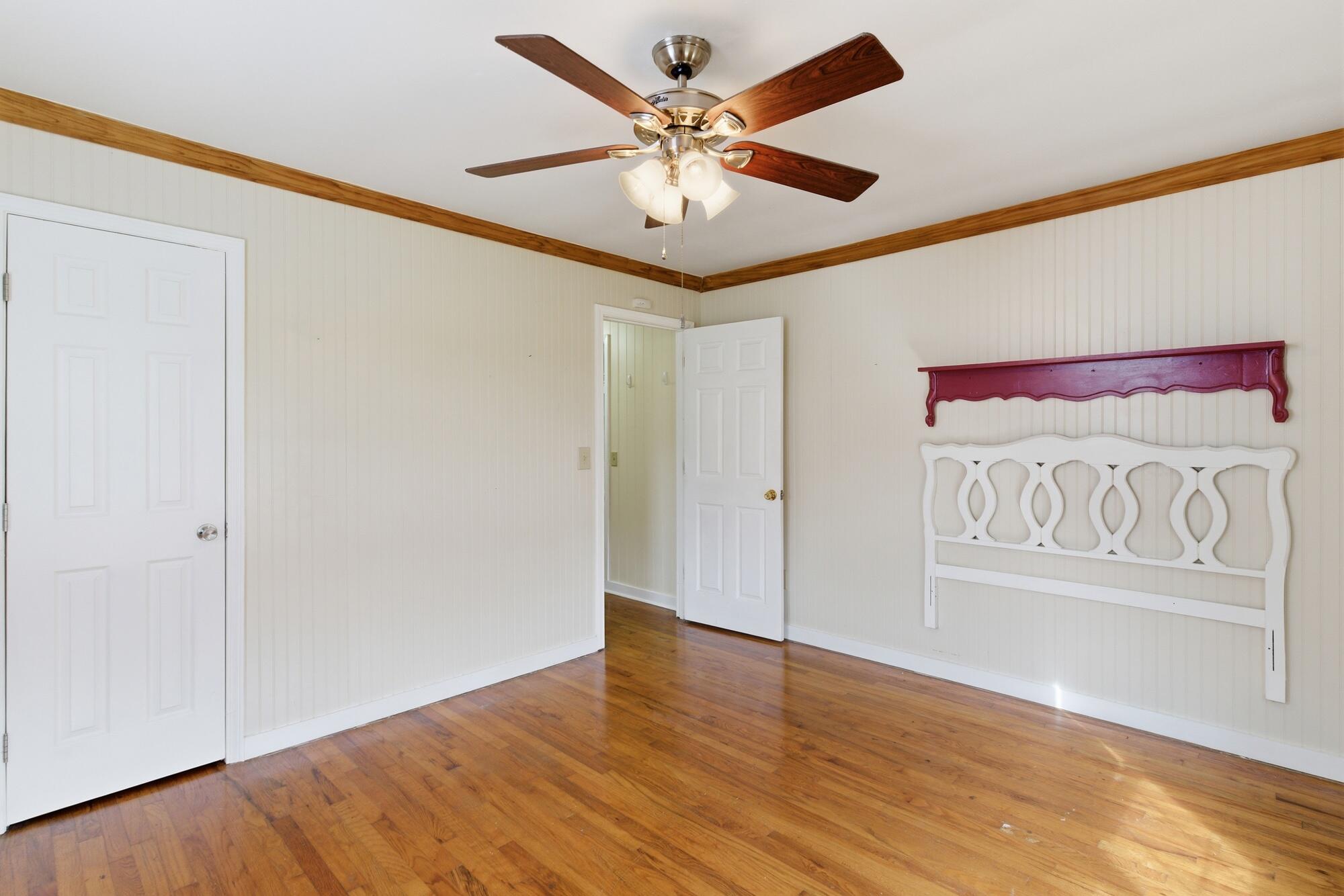 Empty bedroom with hardwood floors, a ceiling fan, and a decorative white headboard mounted on the wall.