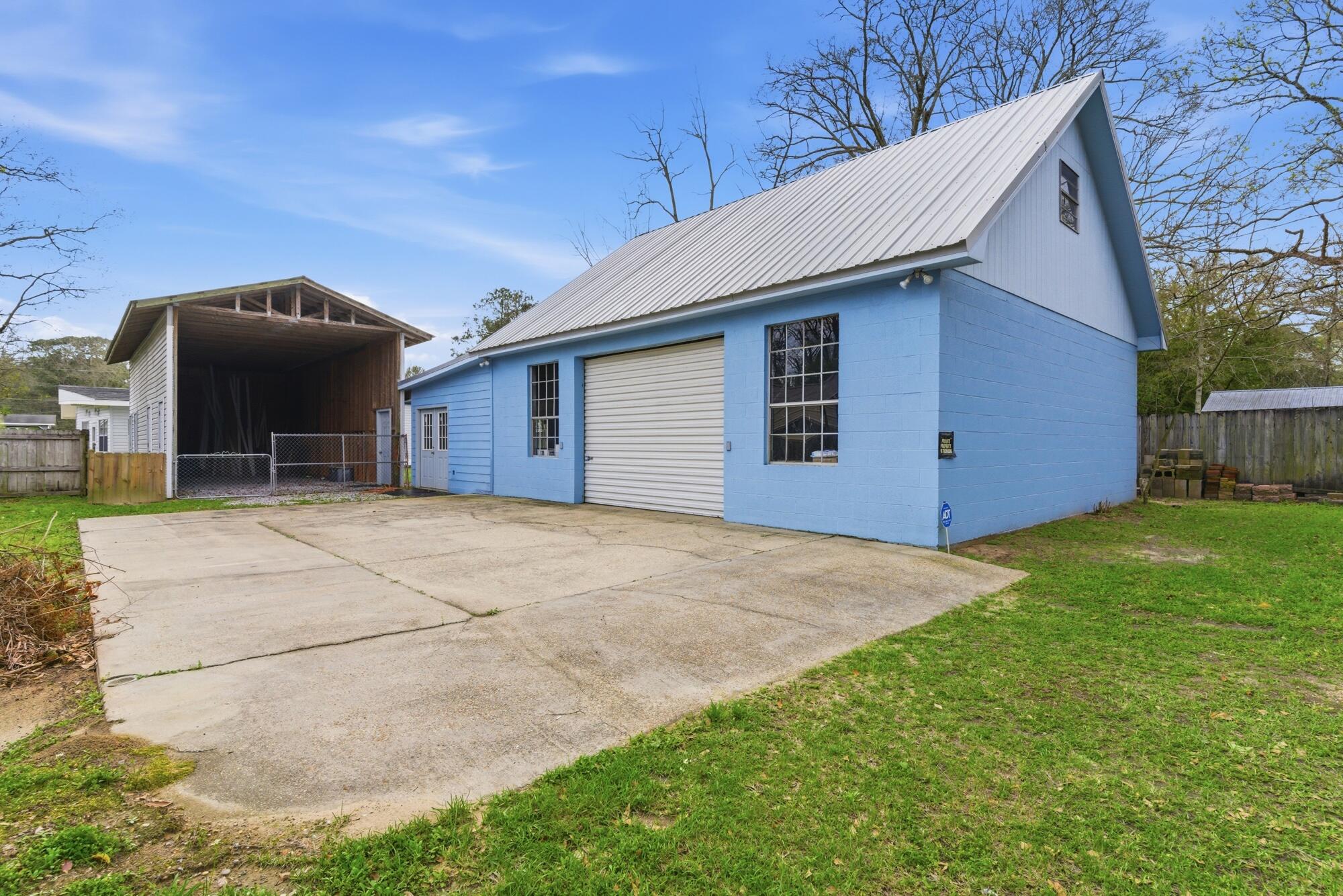 Blue garage with a tall white metal roof and wide concrete driveway beside a covered structure.