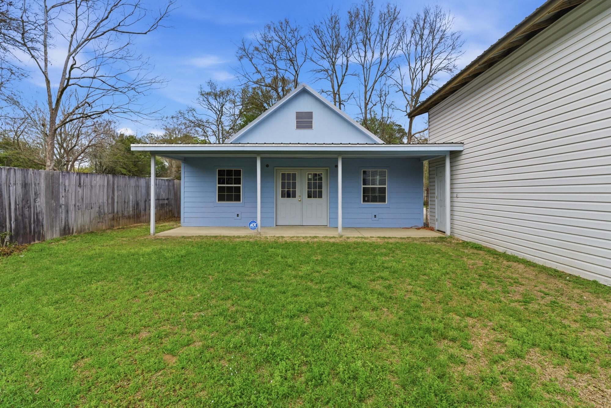 Blue-sided house with a broad covered porch and white double doors, viewed across a grassy fenced yard.