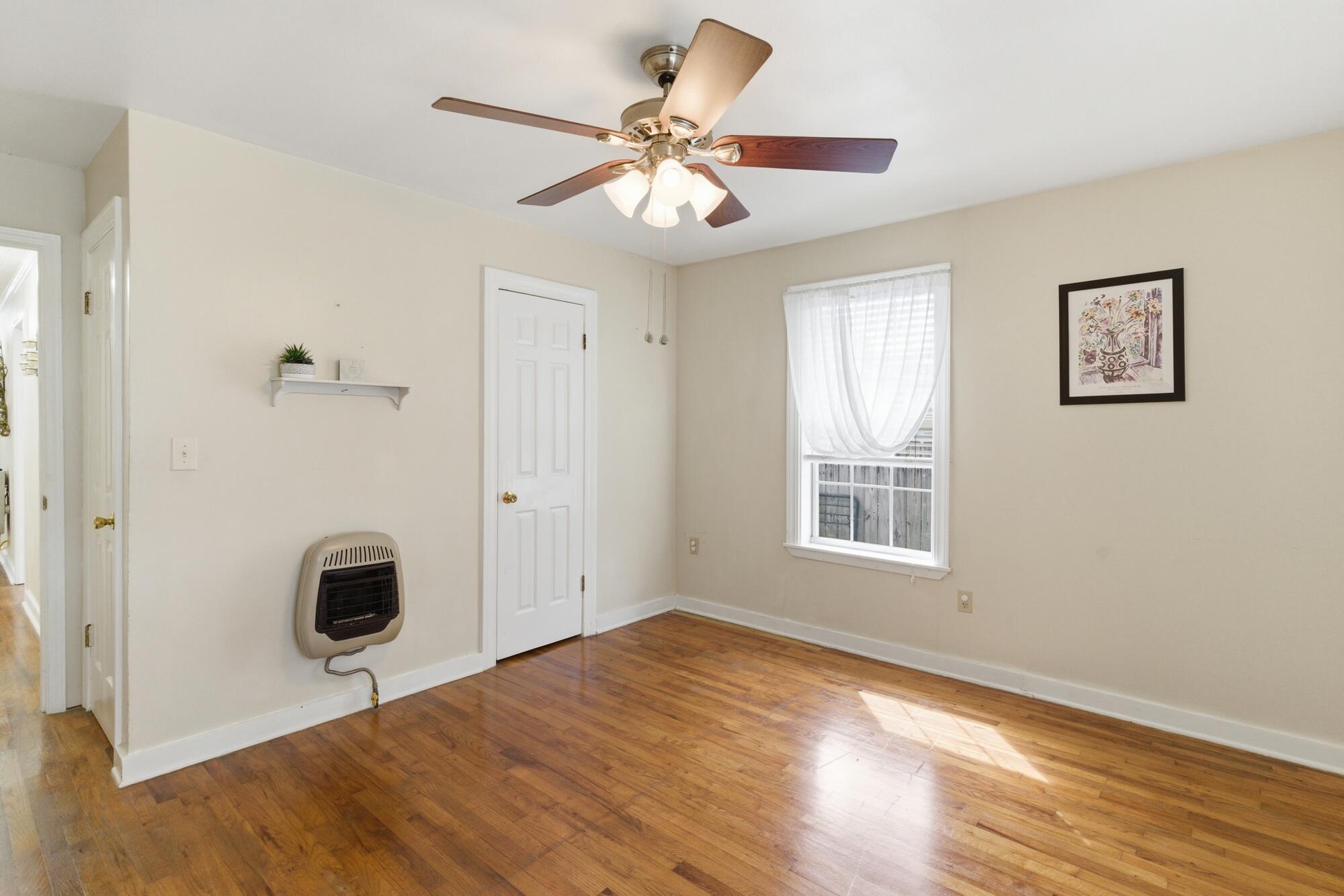 Mostly empty room with hardwood floors, a ceiling fan, wall heater, and curtained window.