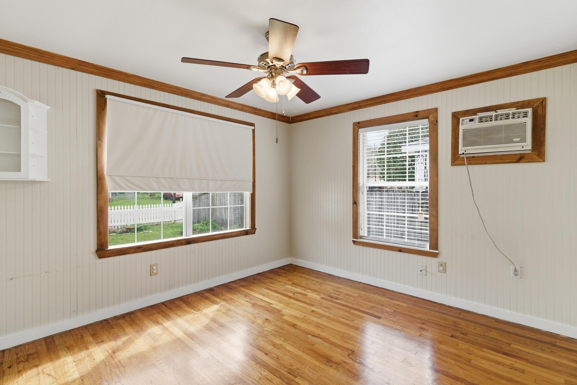 Empty room with hardwood floors, wood-trimmed windows, and a ceiling fan with lights.