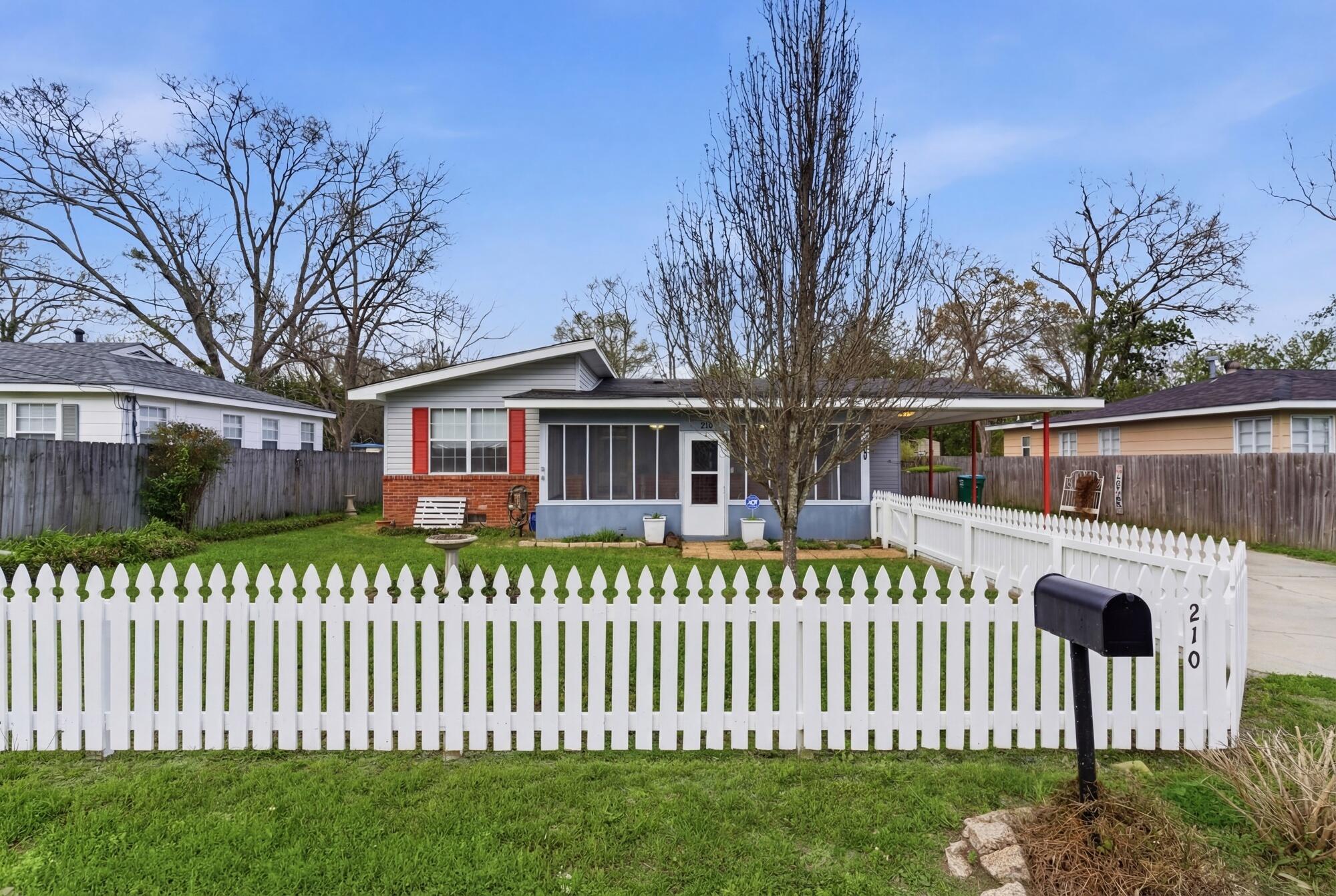 Single-story gray house with red shutters, screened porch, carport, and white picket fence.
