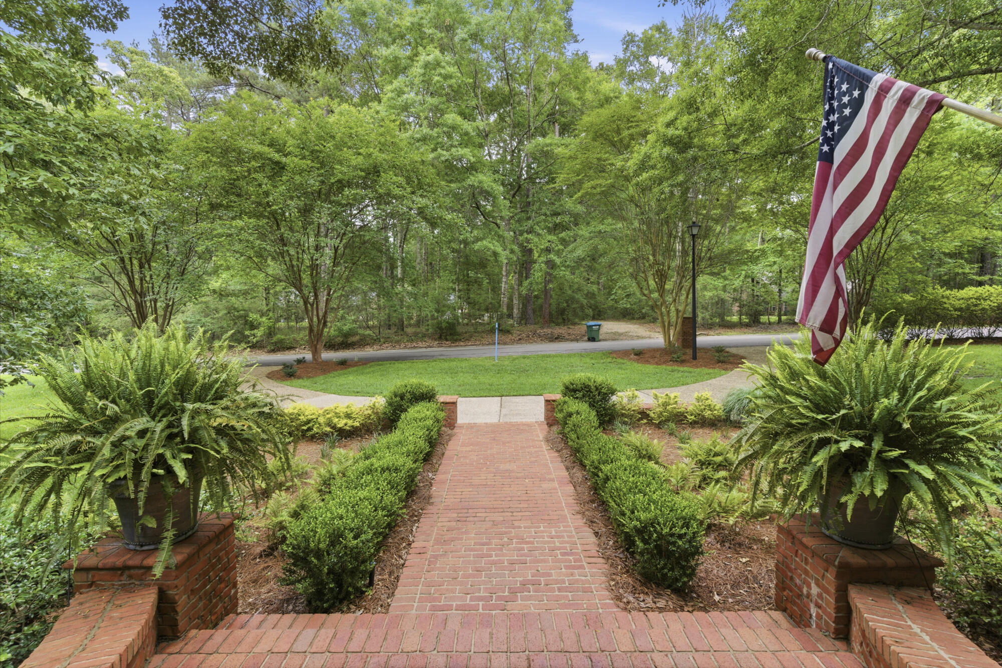 Brick entry walkway bordered by shrubs and potted ferns, with an American flag beside the path.