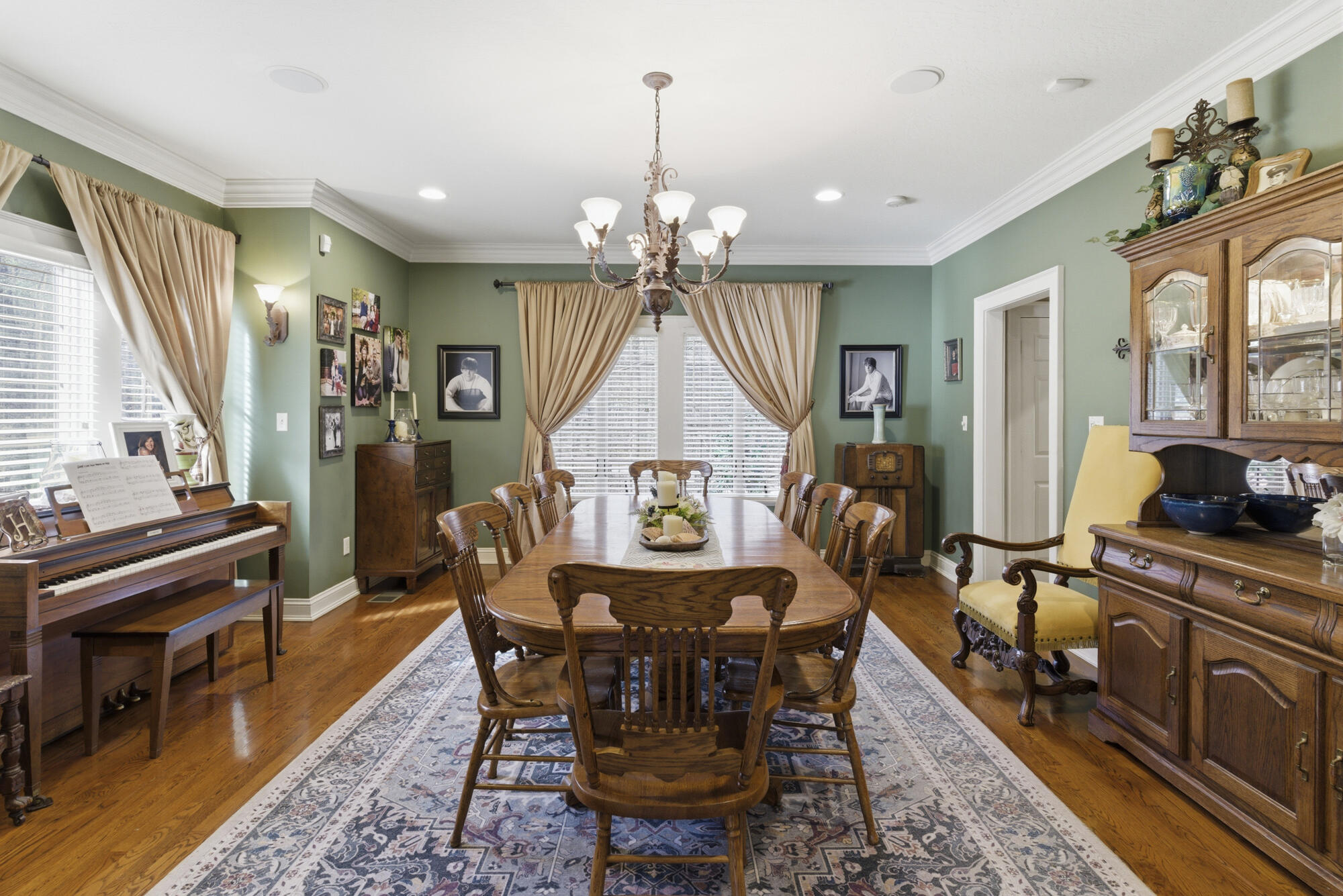 Dining room with a long wooden table, chandelier, piano, and sage-green walls.