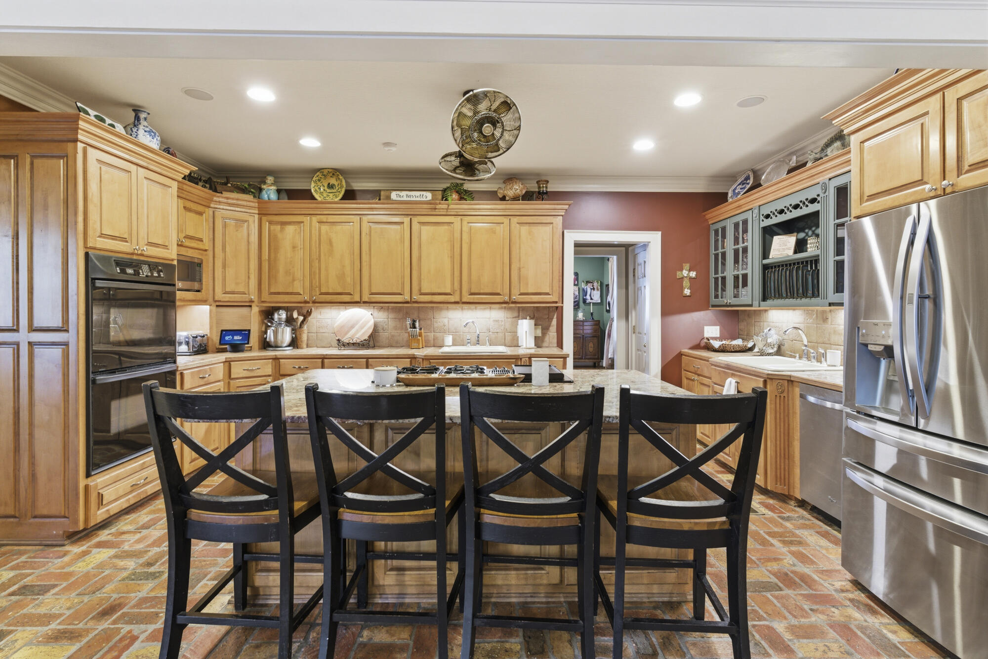 Kitchen with a granite island, four black barstools, honey-colored cabinets, and a brick-tile floor.