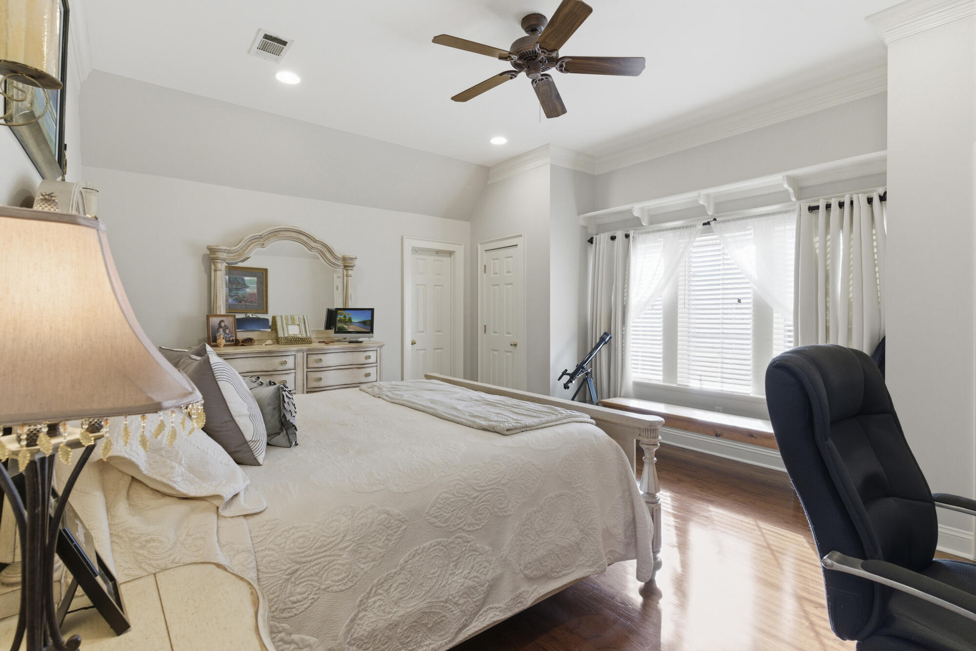 Furnished bedroom with a white quilted bed, hardwood floors, and a bright curtained window.