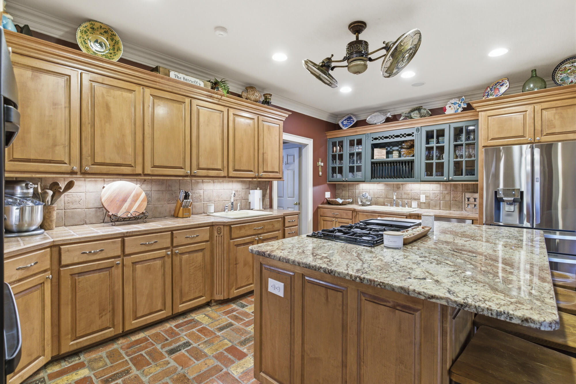 Kitchen with wood cabinetry, blue display cabinets, brick floors, and a granite island with gas cooktop.