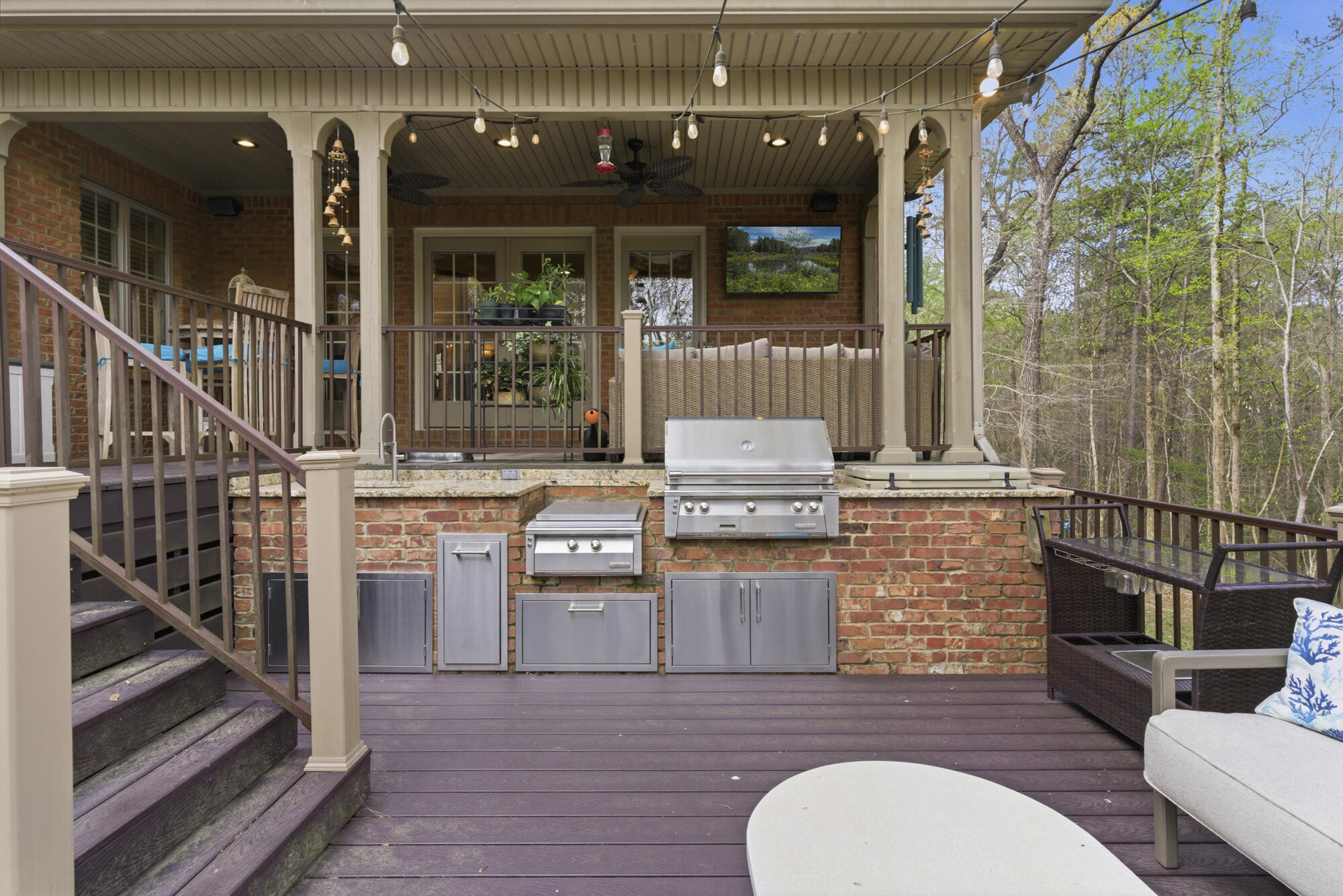 Covered deck with brick outdoor kitchen, stainless steel grills, string lights, and lounge seating beside the woods.