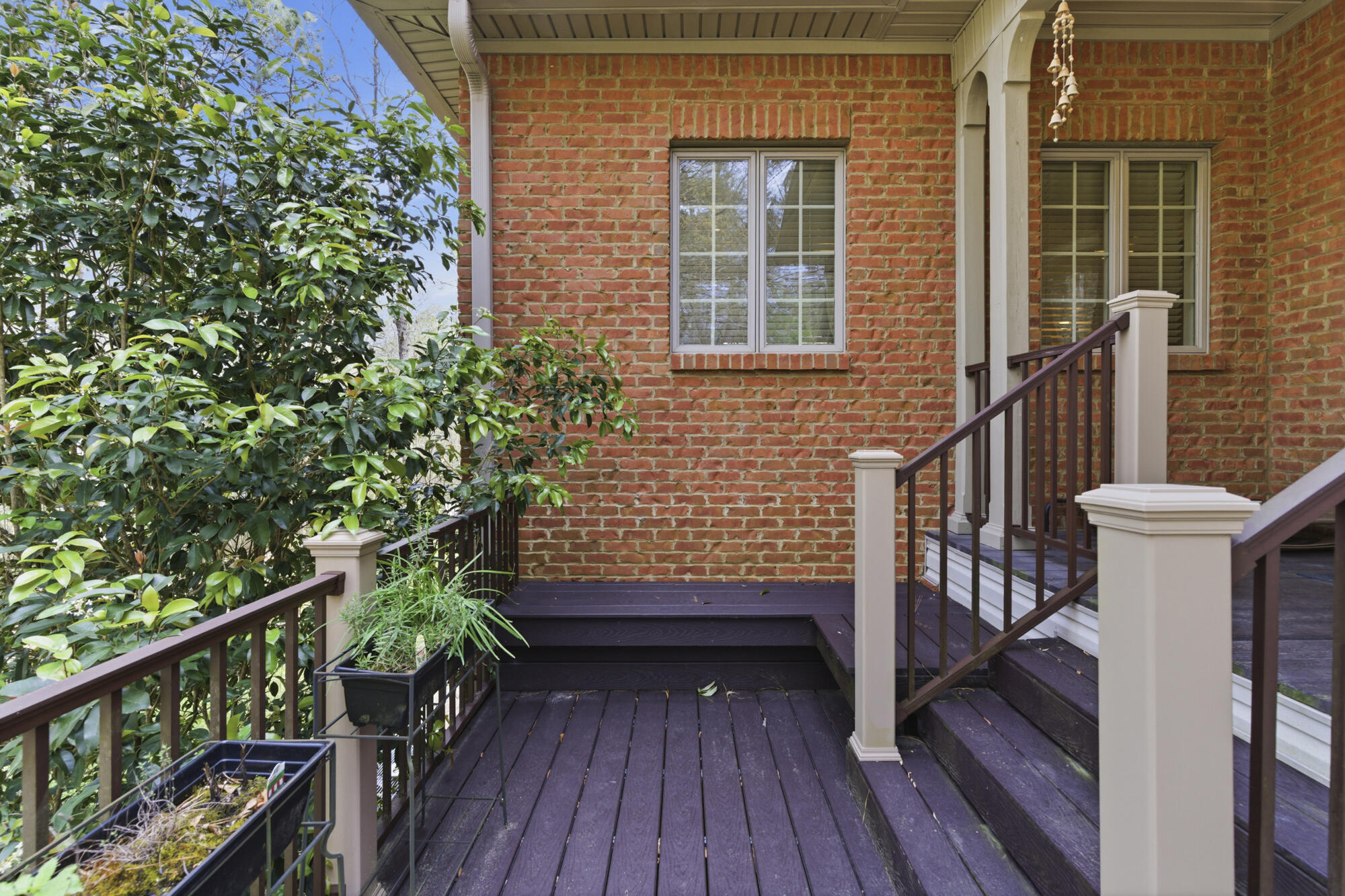 Covered deck beside a red brick home, with dark wood railings and dense greenery.