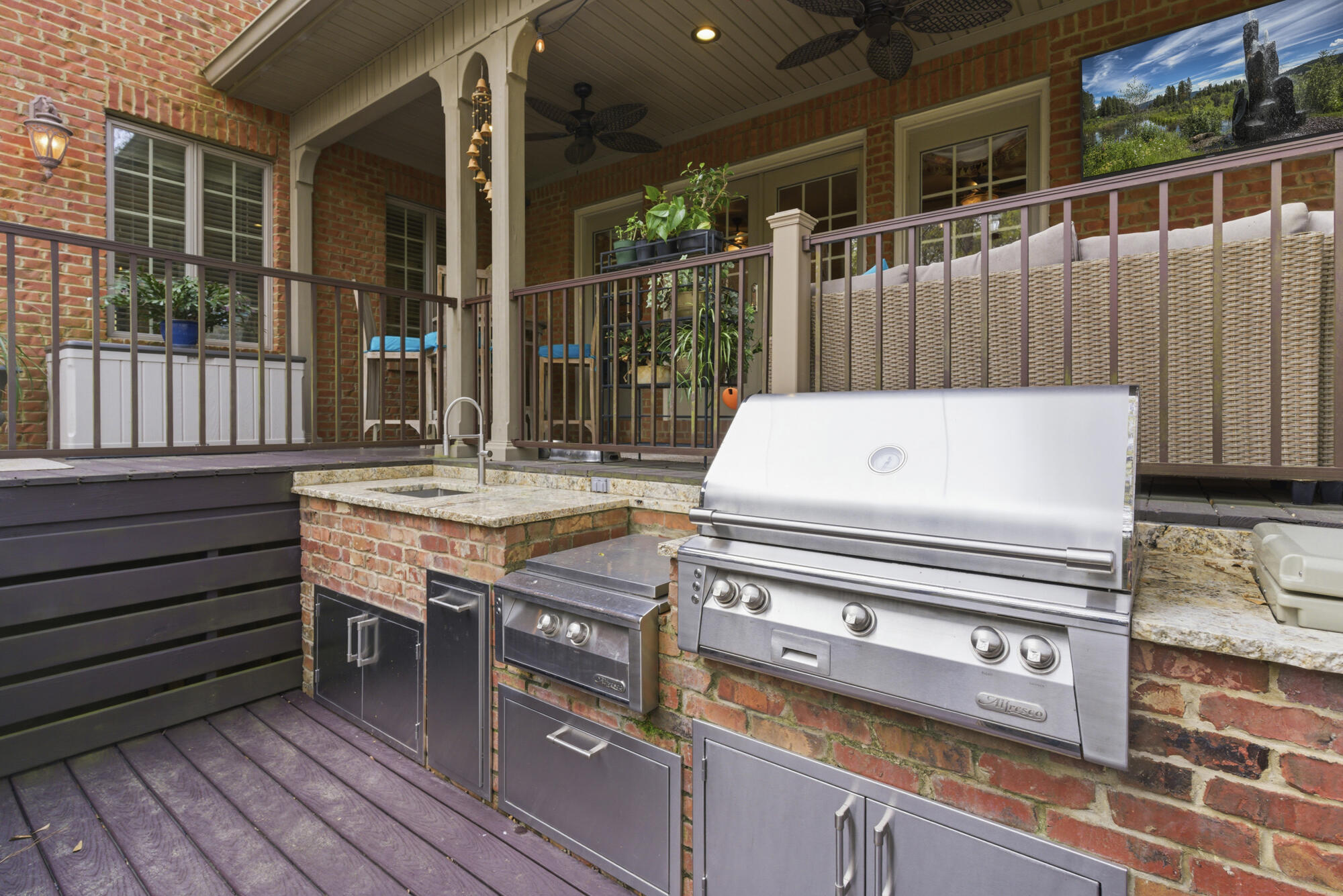 Covered outdoor kitchen with a stainless-steel grill, brick counters, sink, and deck seating.