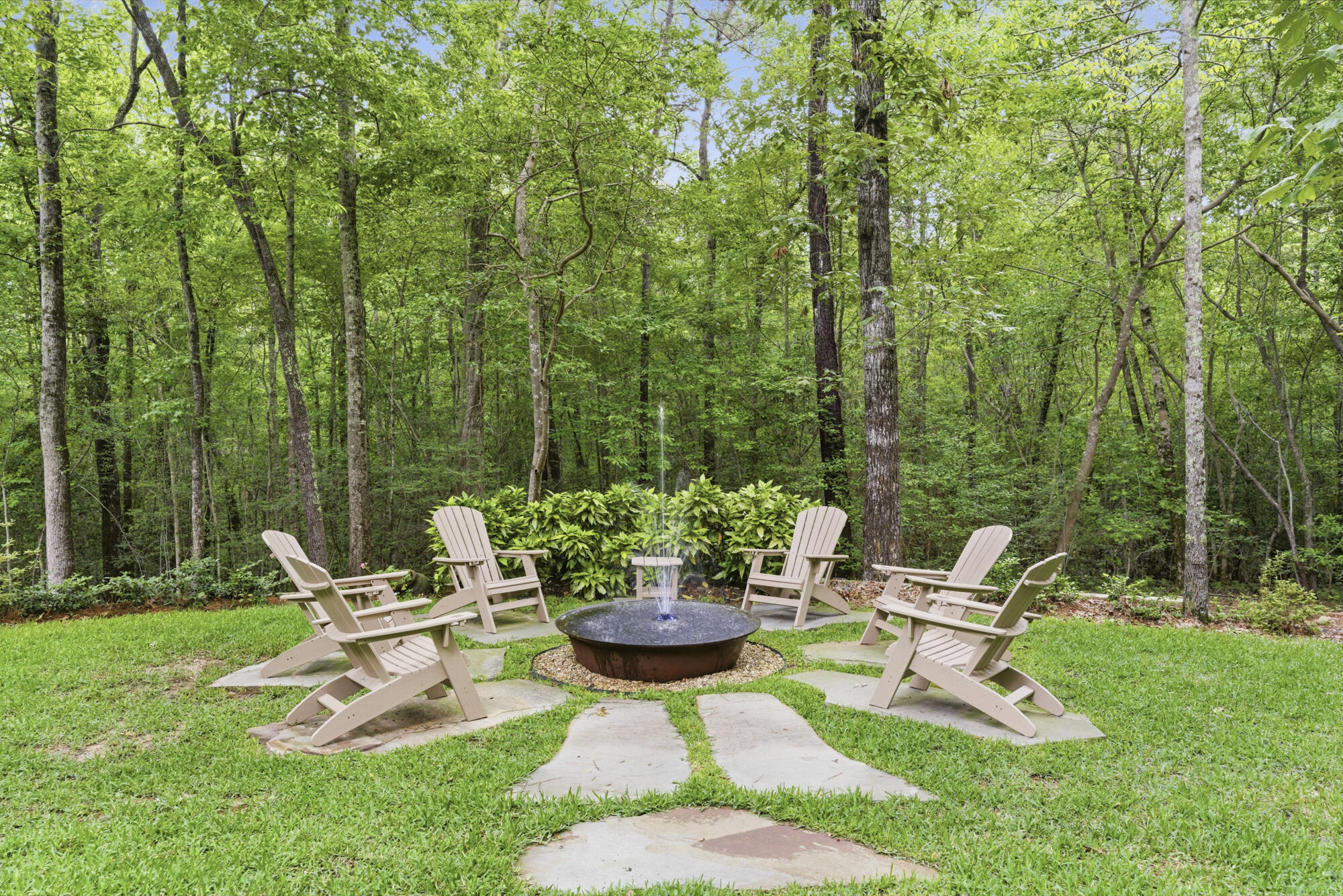 Backyard patio with Adirondack chairs around a bubbling water feature, bordered by green woods.