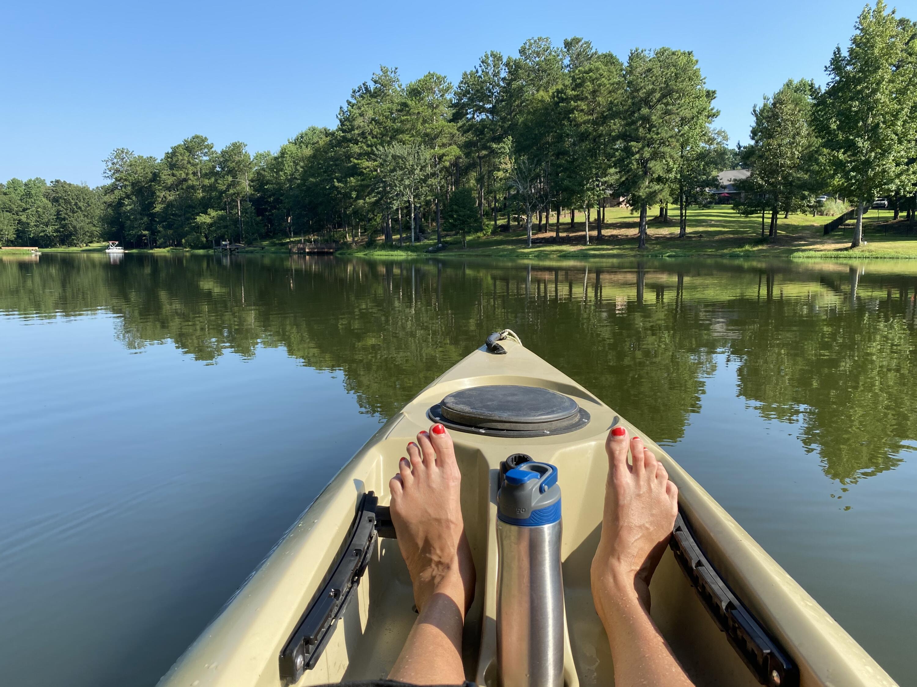Kayak on a calm, tree-lined lake, viewed from the seat with bare feet in the foreground.