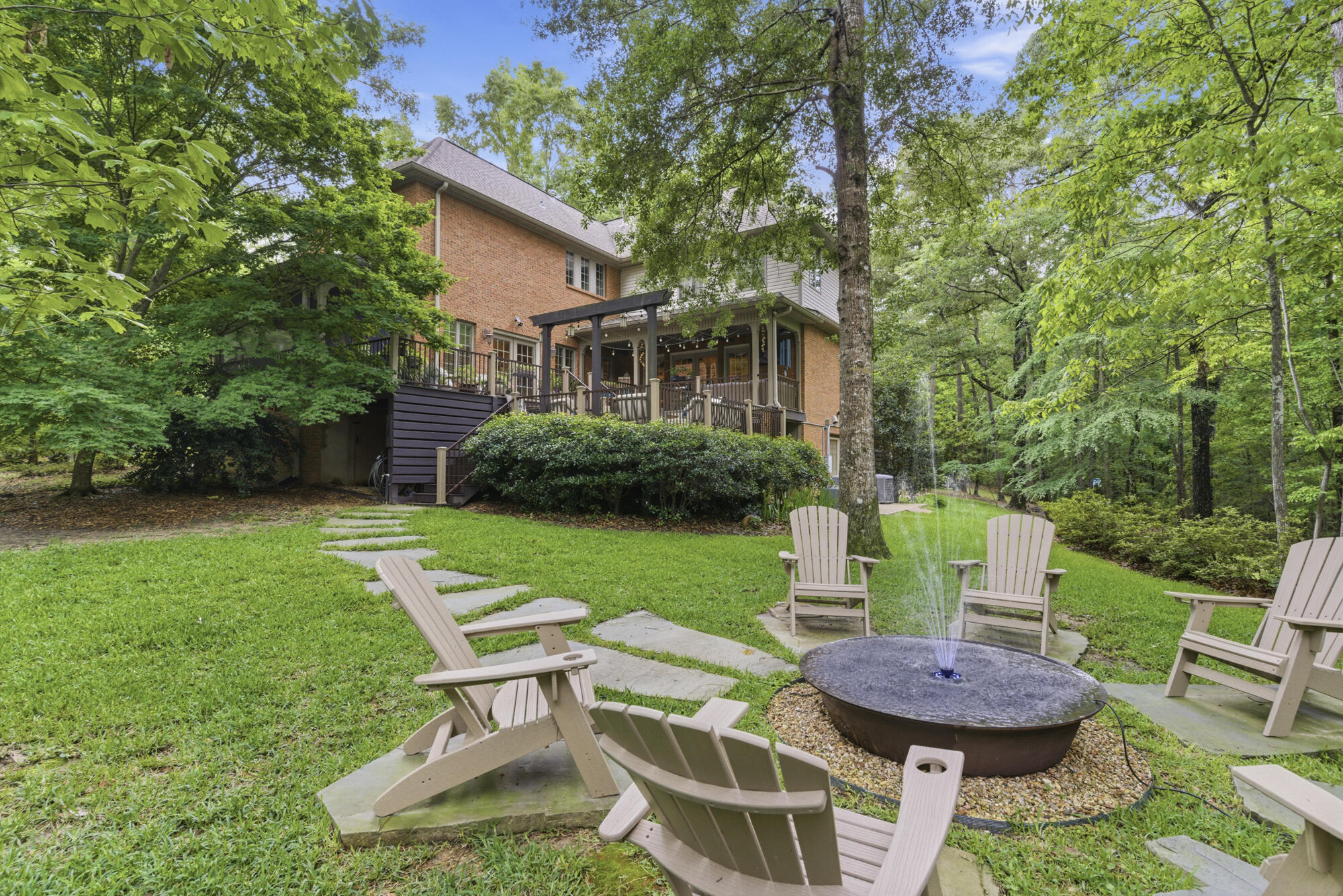 Brick home with a raised deck behind a lawn and a fountain patio surrounded by trees.