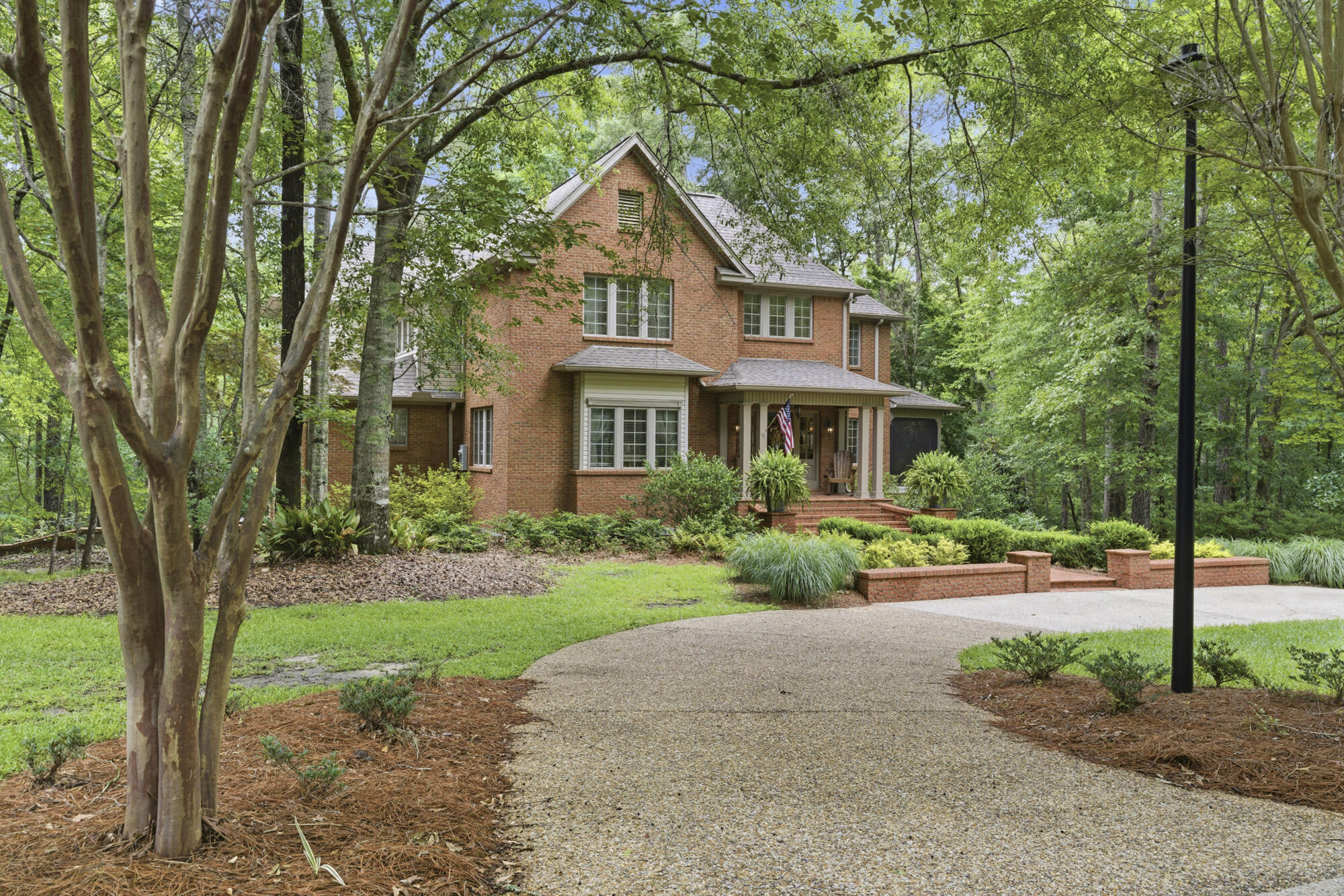 Two-story brick house with a covered porch, landscaped yard, and curved gravel driveway beneath mature trees.