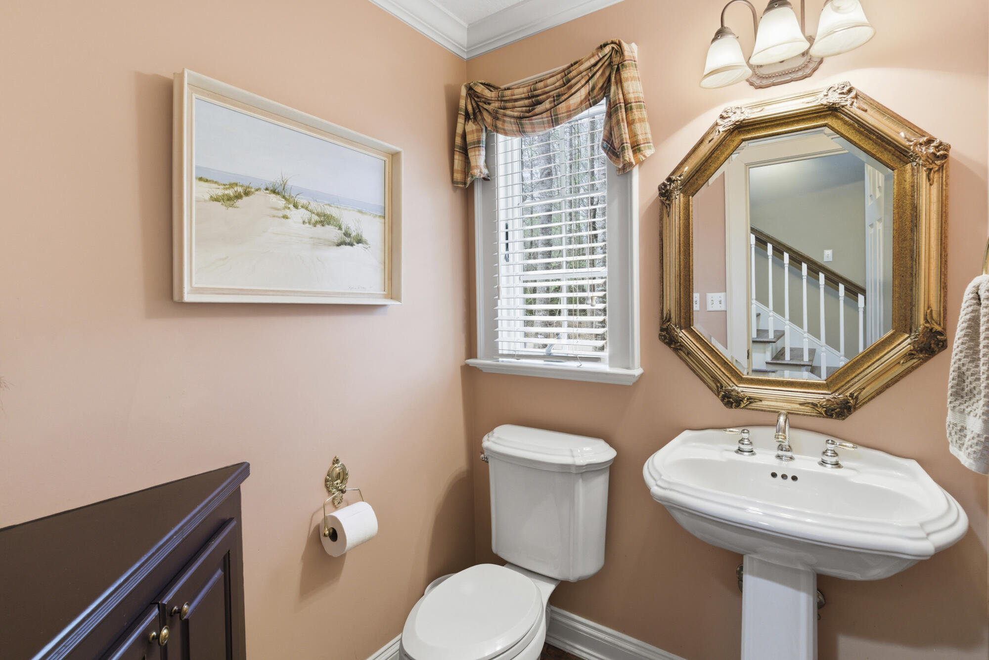 Bathroom with a white pedestal sink, ornate gold mirror, shuttered window, and peach walls.