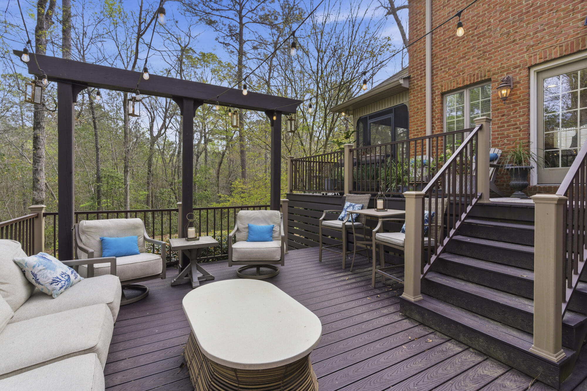 Furnished deck with a lit pergola, lounge seating, and wooded views beside a brick home.