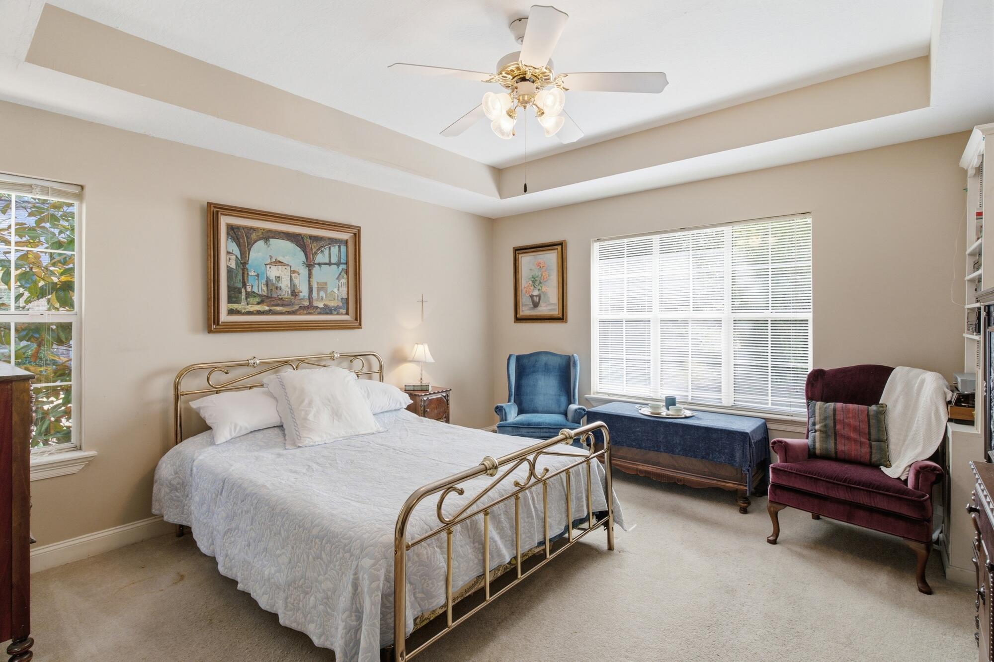 Bedroom with brass bed, tray ceiling, and blue and burgundy chairs beside a large window.
