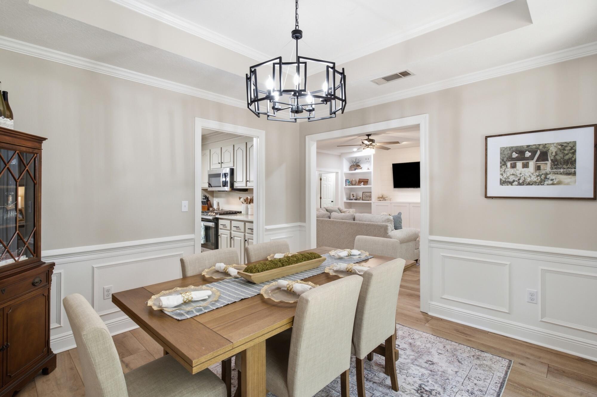 Dining room with a wooden table, cream chairs, and a geometric black chandelier, opening to the kitchen and living room.