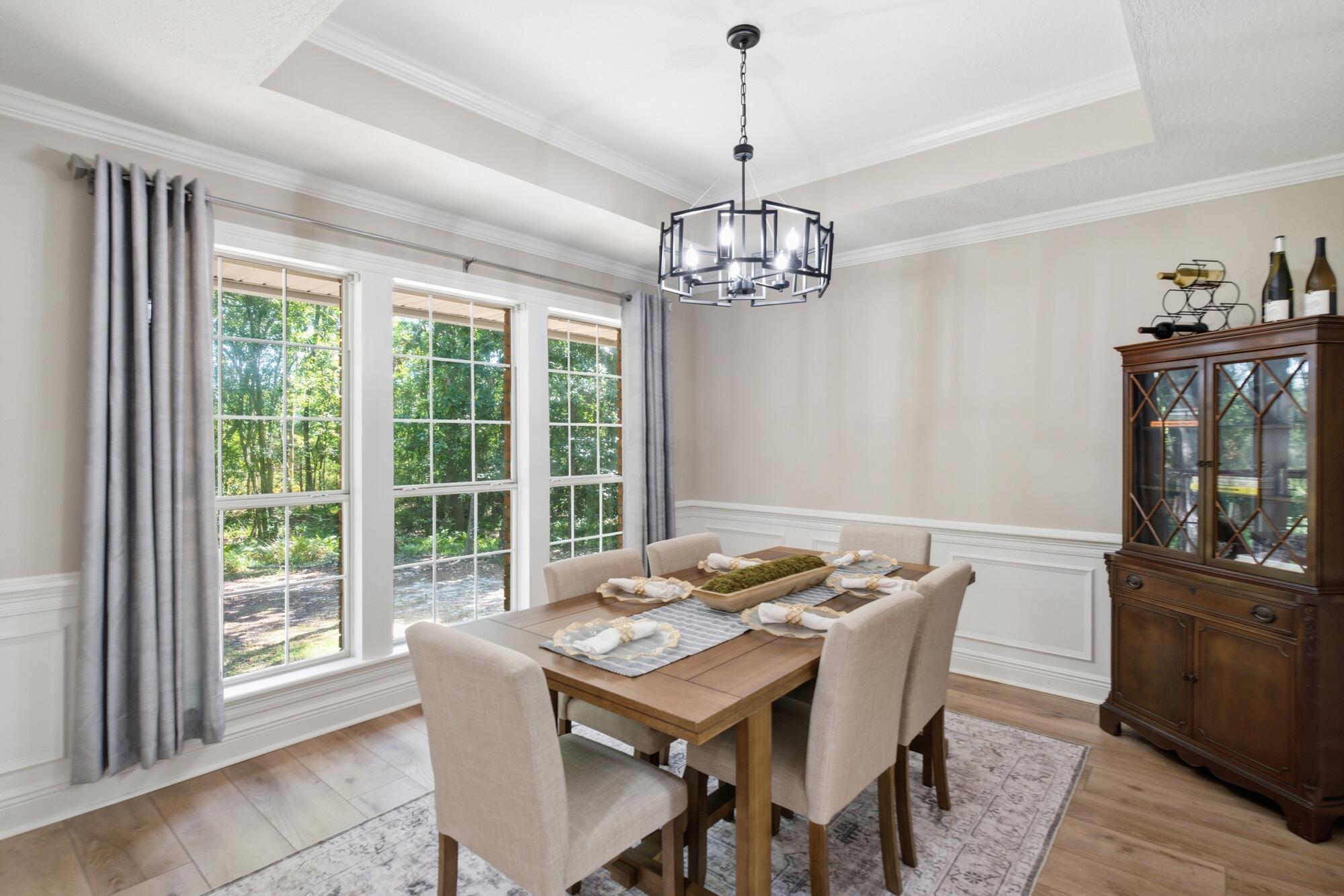 Dining room with a wooden table, geometric chandelier, and tall windows overlooking trees.
