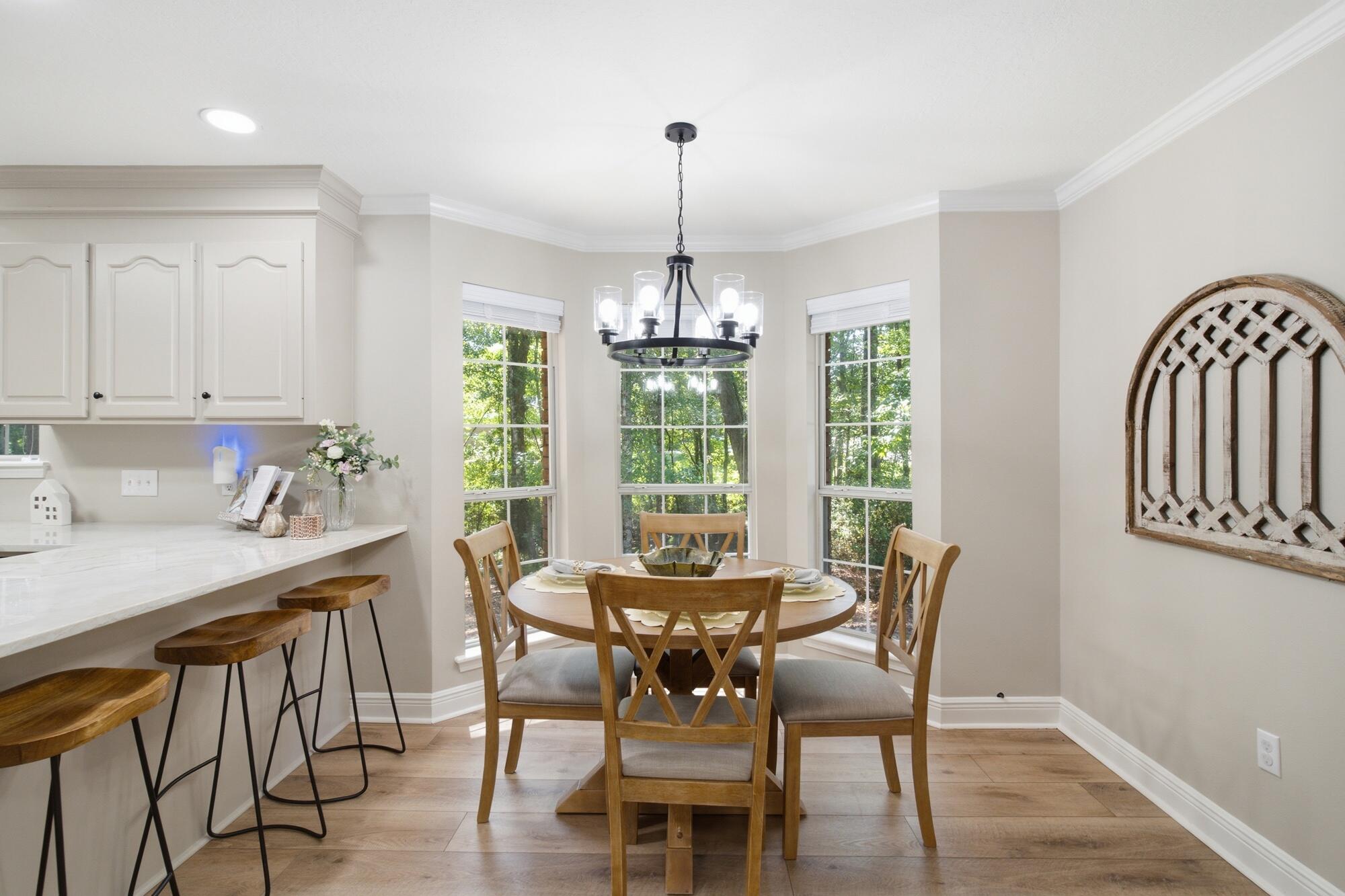 Dining area with a round wooden table, black chandelier, and three tall windows overlooking trees.