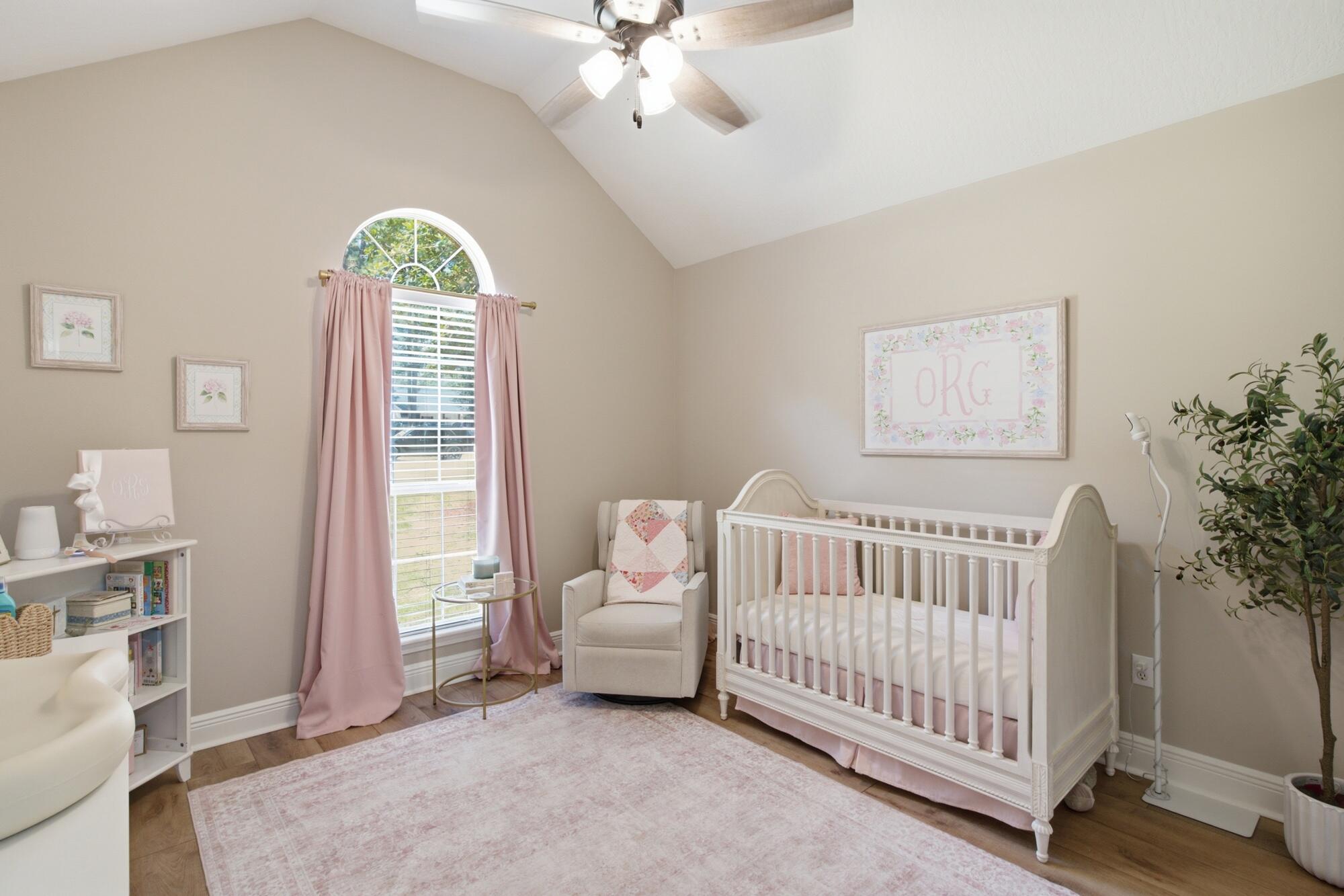 Nursery with a white crib, glider, pink curtains, and a tall arched window.