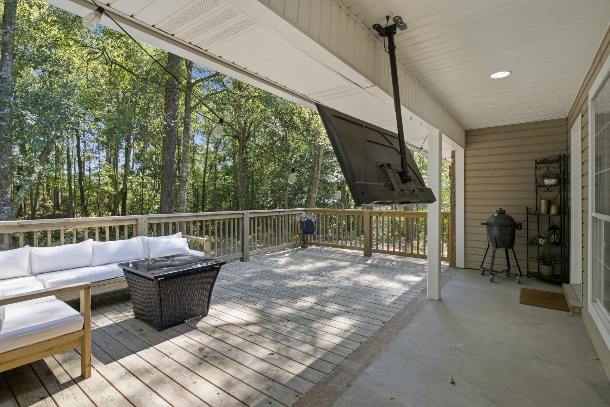 Covered wooden deck with white seating, mounted television, and a wooded view.
