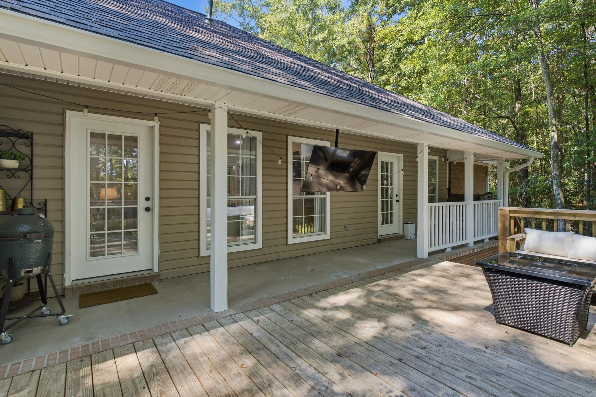 Covered back porch and deck with a wall-mounted television, outdoor seating, and trees beyond.