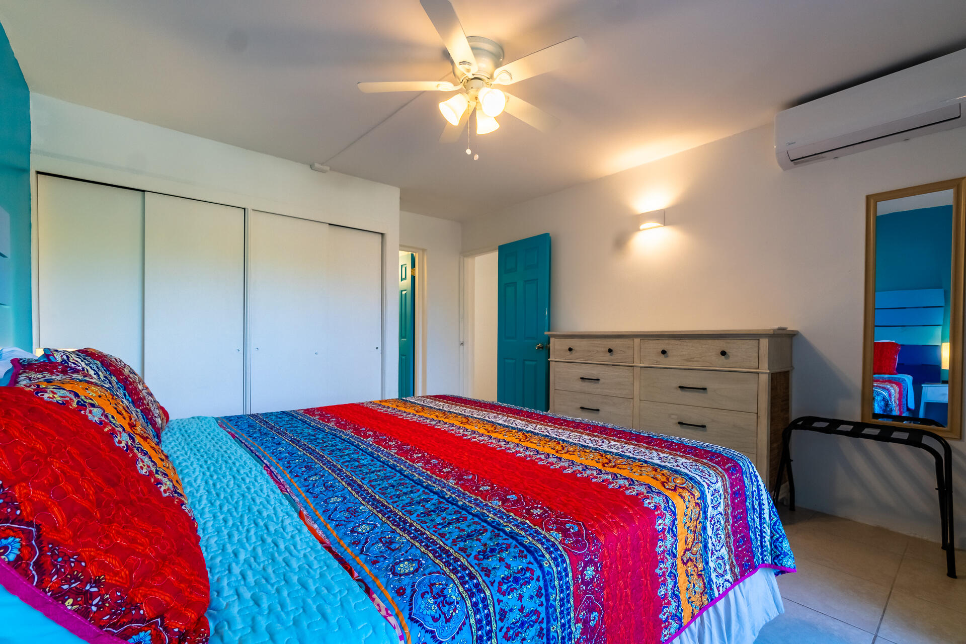 Bedroom with vivid patterned bedding, white sliding closets, a wooden dresser, and turquoise door.