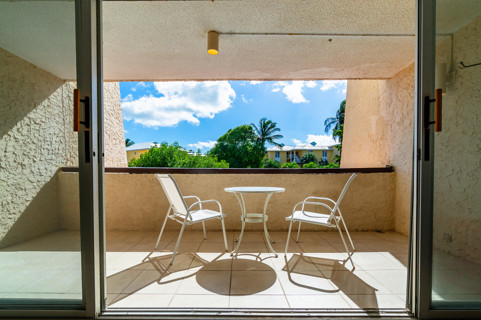 Tiled balcony with two white chairs and a table overlooking palms and neighboring residences.