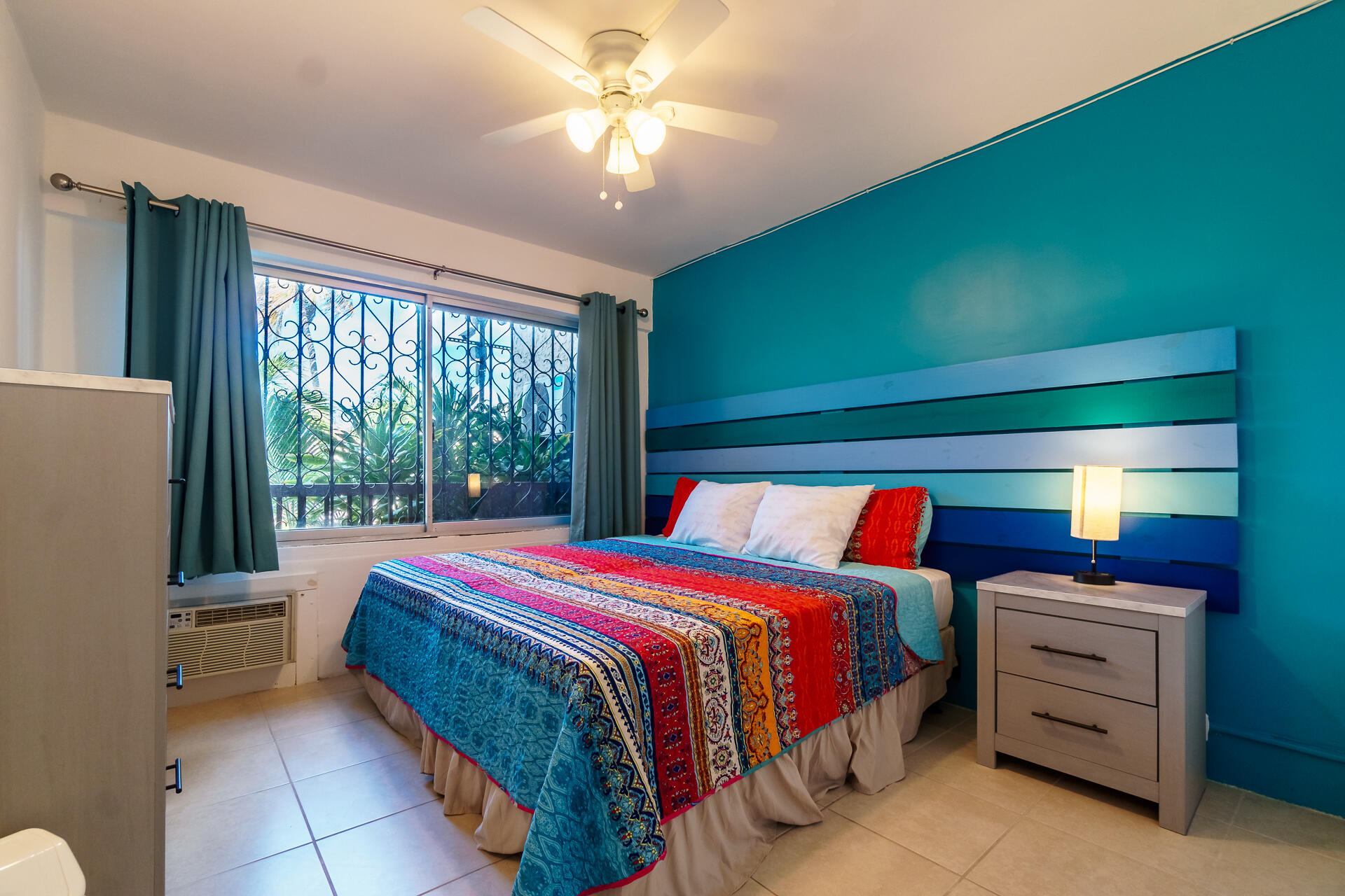 Furnished bedroom with colorful bedding, a teal accent wall, and a window overlooking tropical greenery.