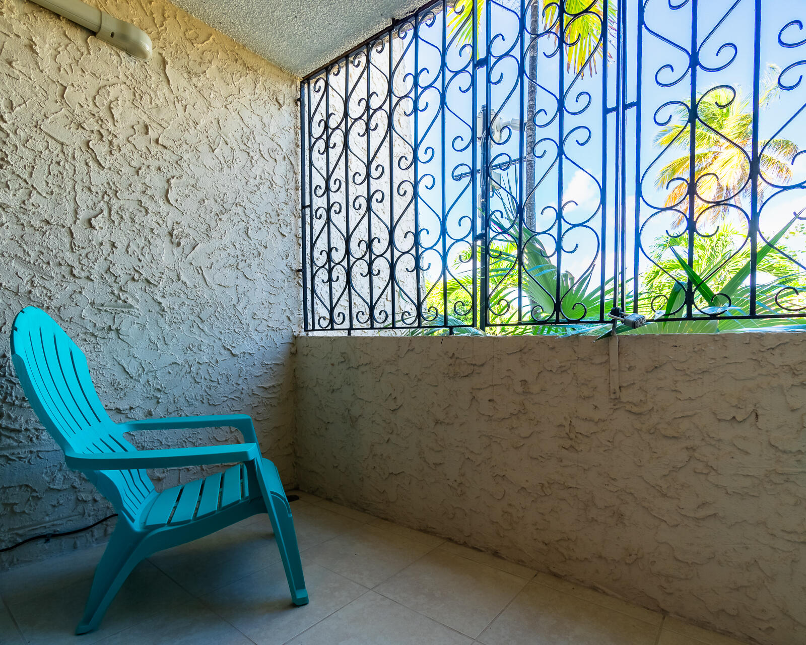 Tiled patio with a turquoise chair, ornate metal grilles, and palm foliage outside.