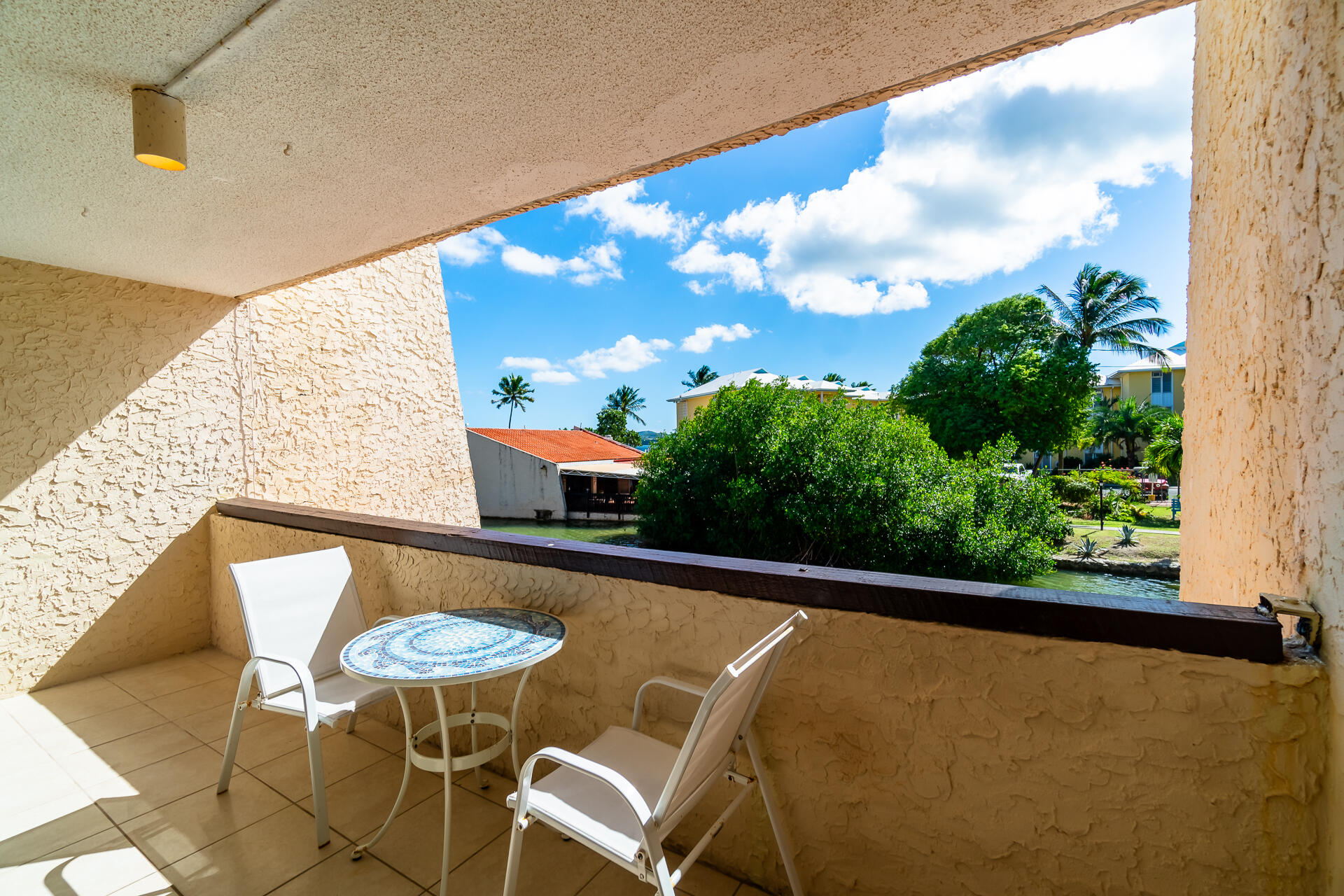 Covered balcony with white chairs and mosaic table overlooking a canal and tropical greenery.