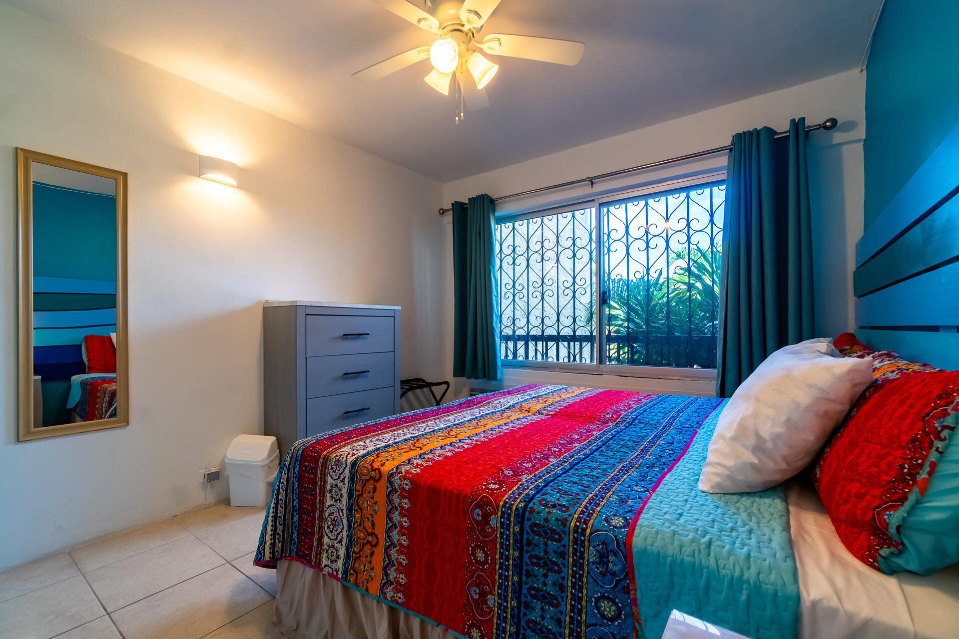Bedroom with a vivid patterned bedspread, teal curtains, and a barred window overlooking plants.