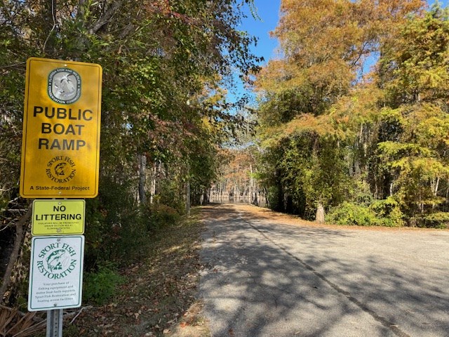 Tree-lined road to a public boat ramp, with autumn foliage and roadside signs.
