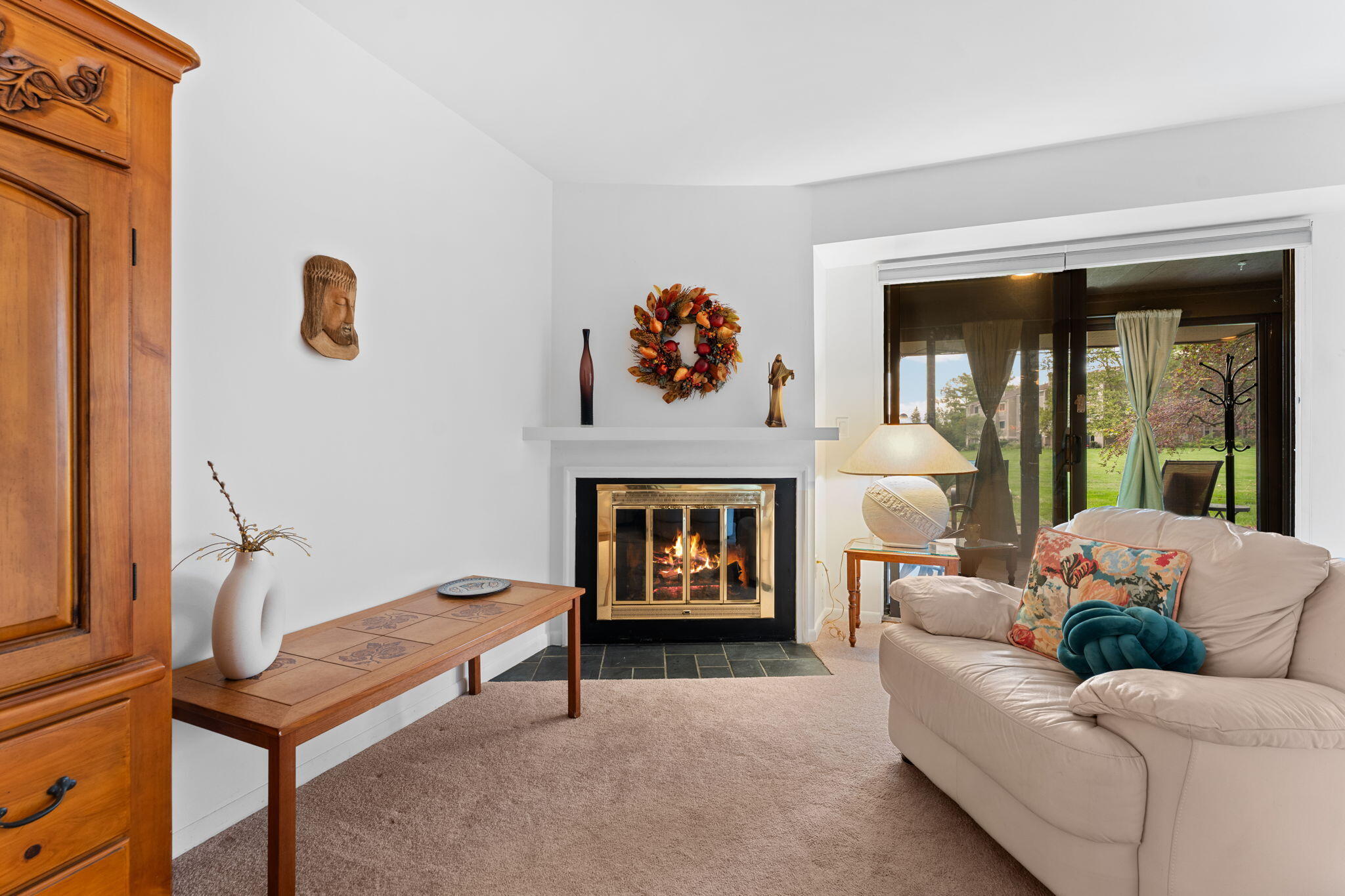 Living room with a lit fireplace, beige sofa, and sliding glass doors overlooking a lawn.