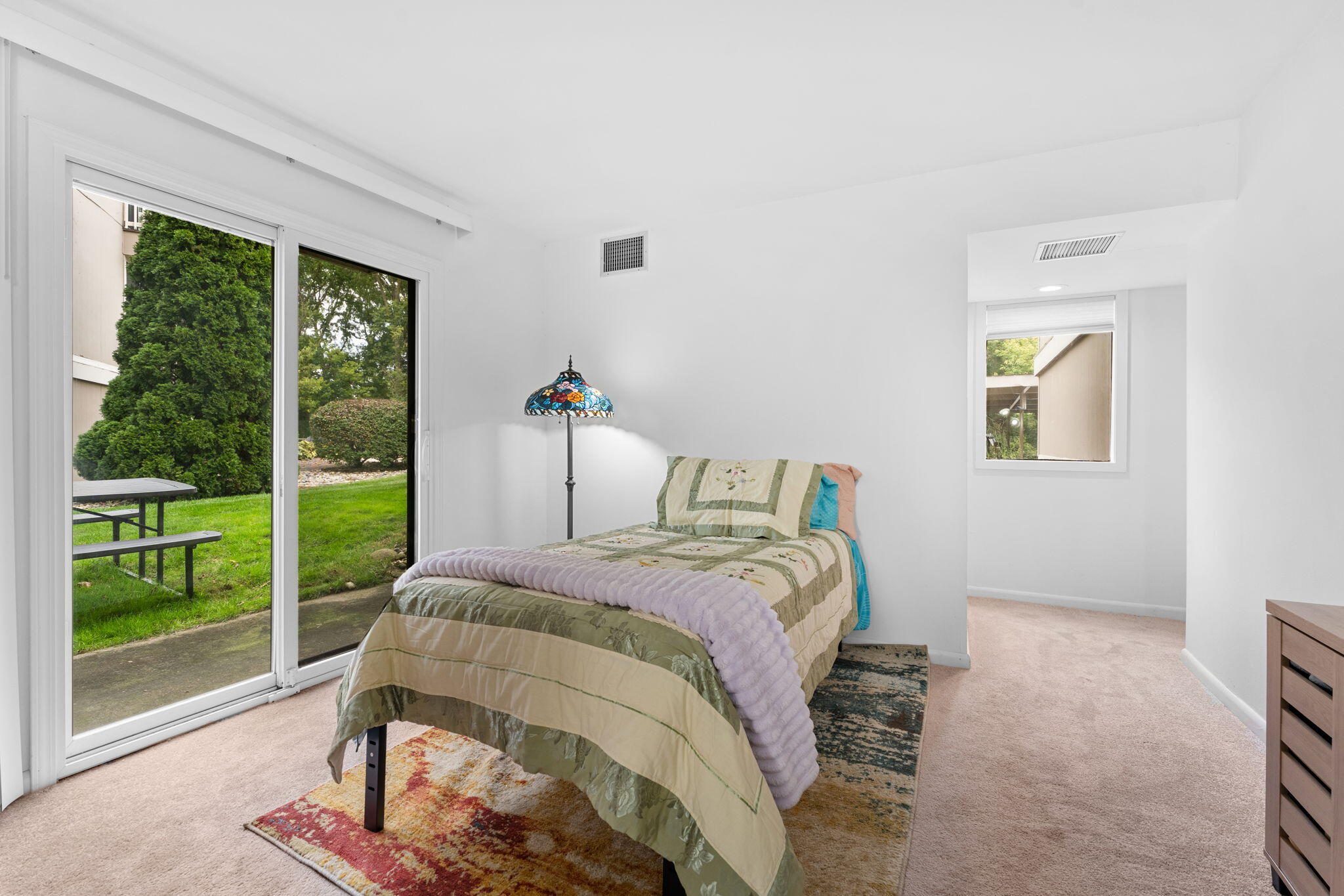 Bedroom with a floral quilted bed beside a sliding glass door overlooking a lawn.