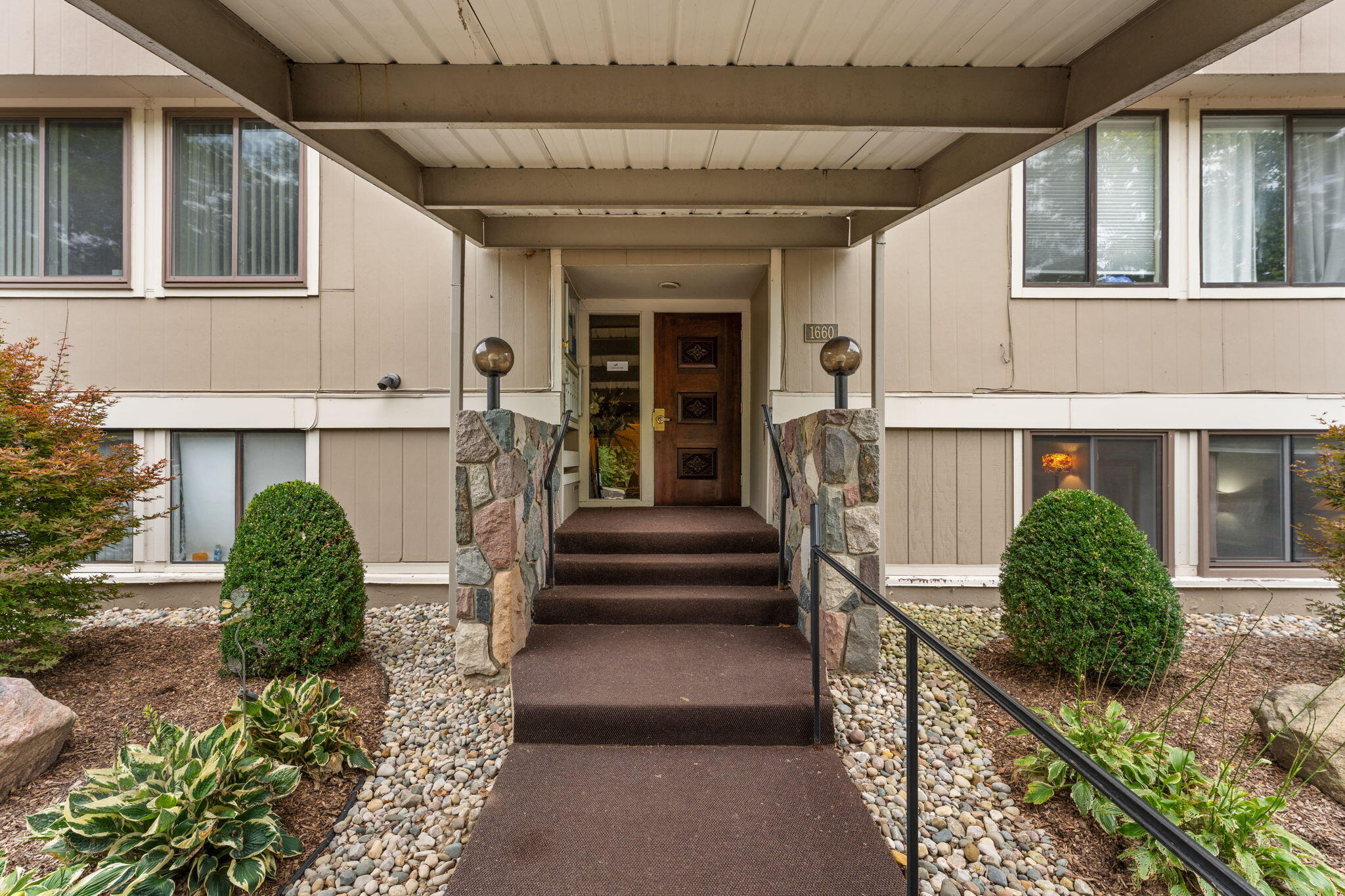 Covered residence entrance with carpeted steps, stone sidewalls, black handrails, and landscaped beds.