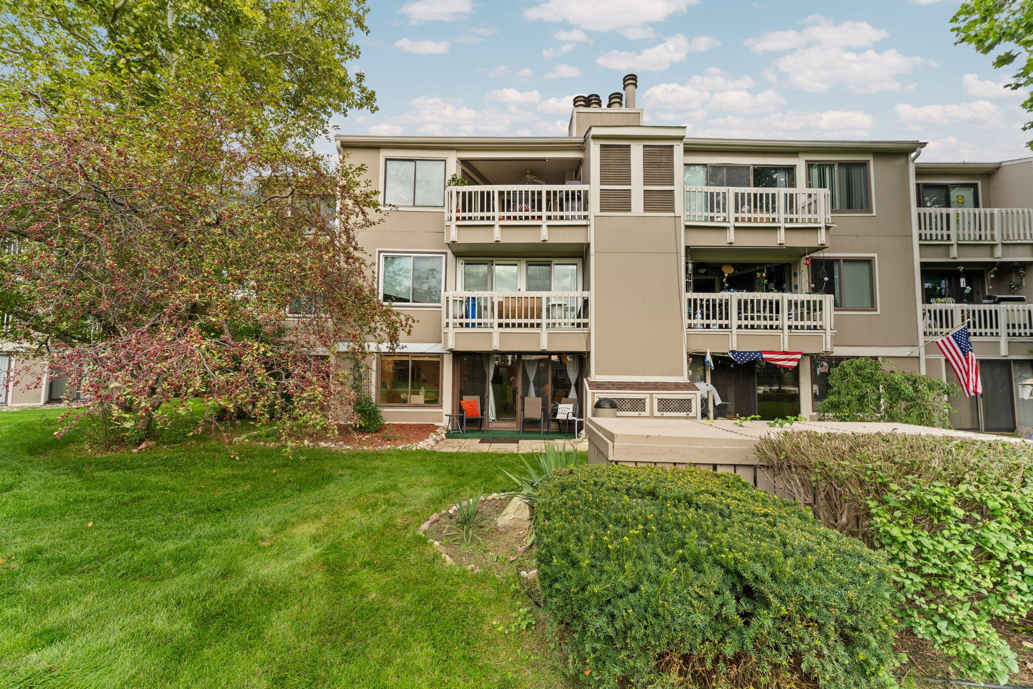 Beige condominium building with multiple balconies overlooking a landscaped lawn and shrubs.