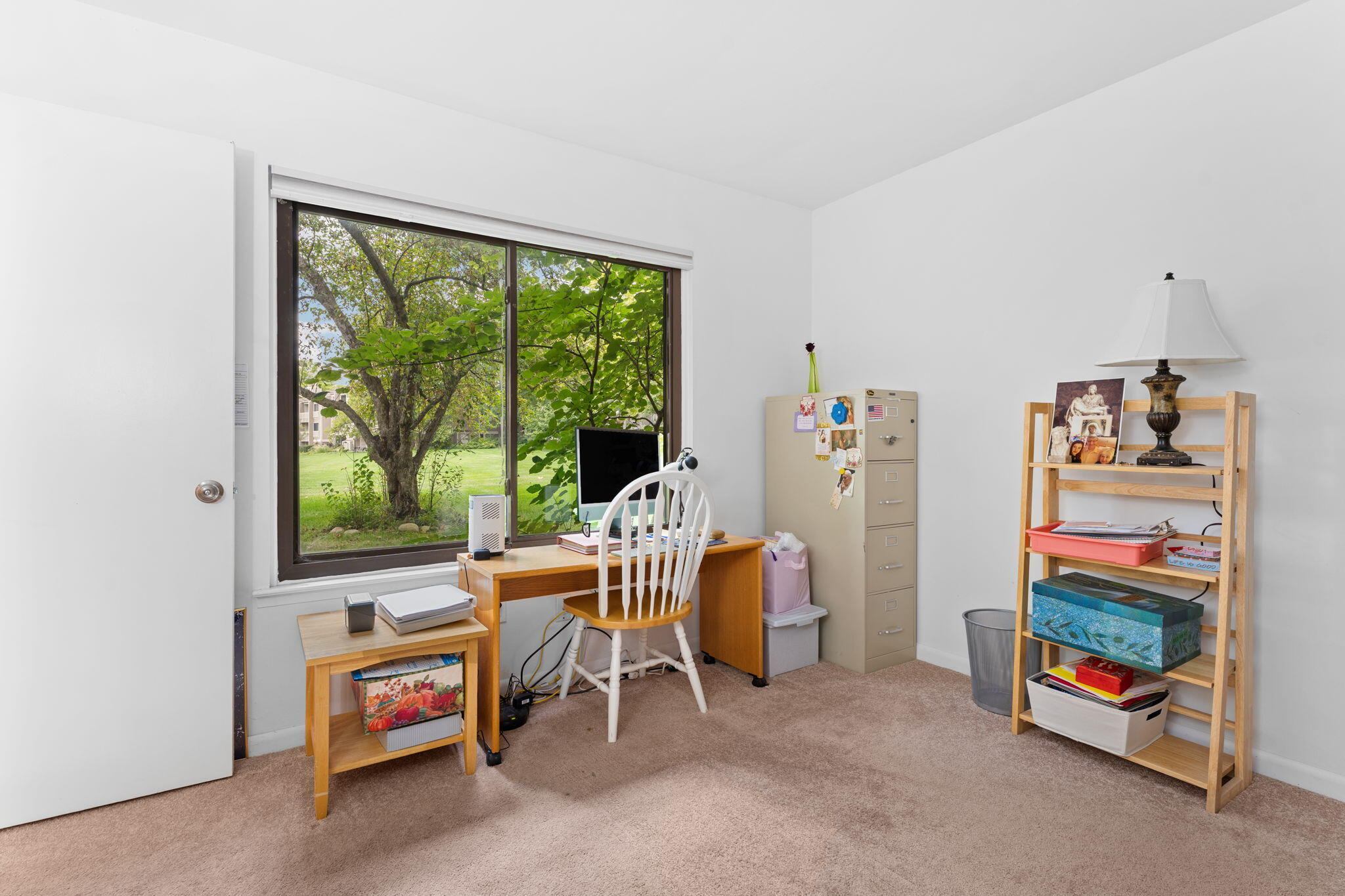Home office with a wooden desk and chair beside a large window overlooking leafy trees.