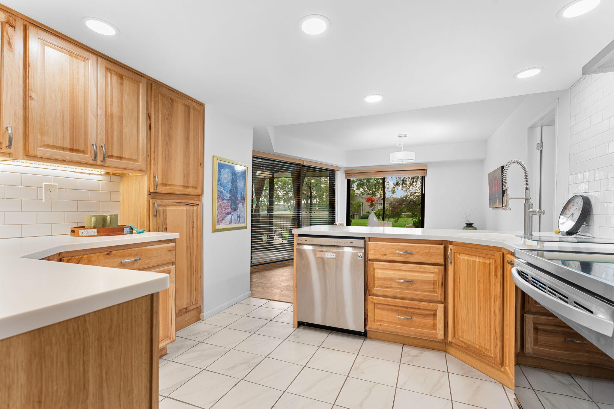 Kitchen with honey-colored wood cabinets, a stainless steel dishwasher, and garden views through large windows.