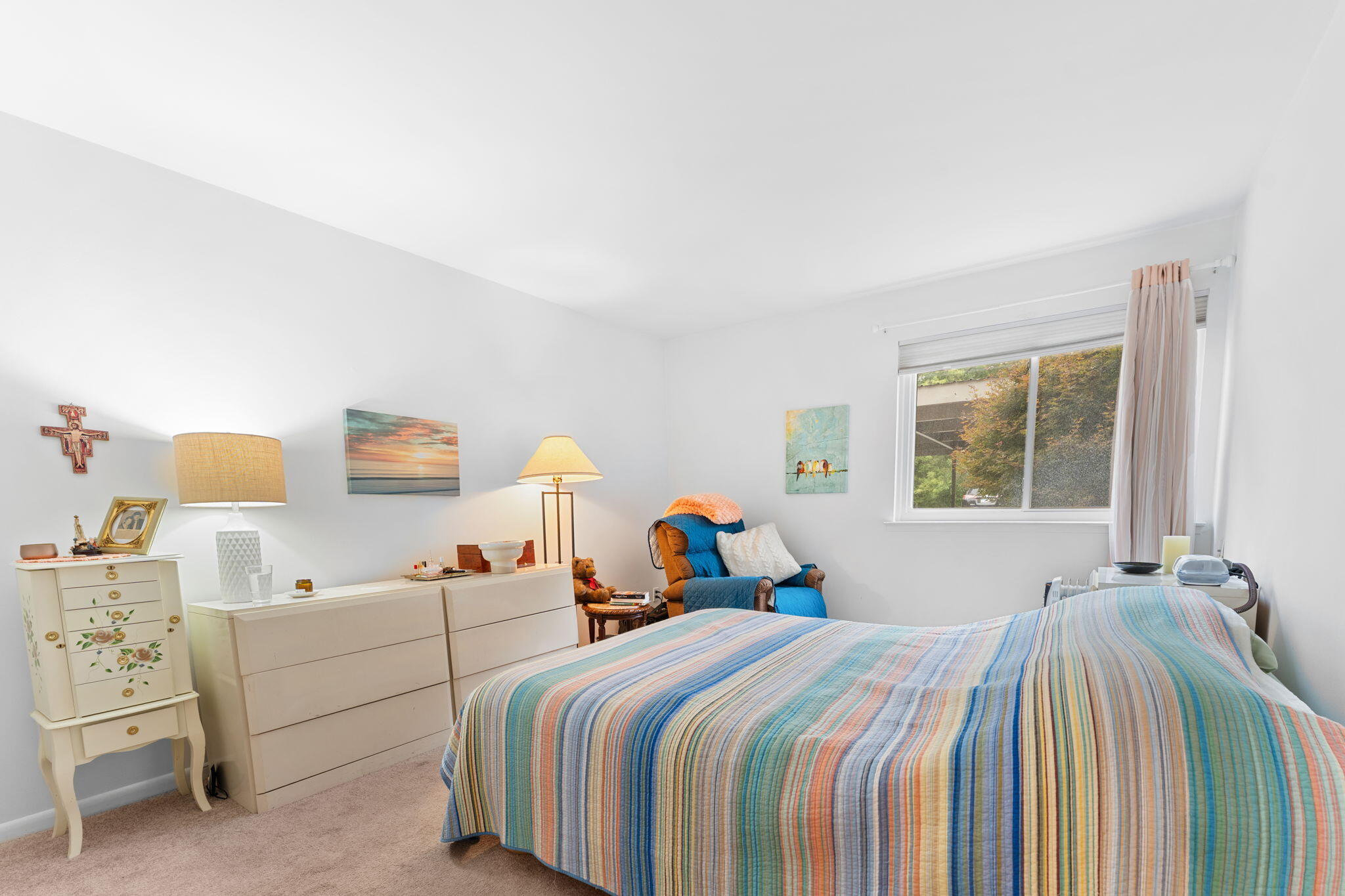 Furnished bedroom with a striped quilt, white dressers, and a blue armchair by the window.