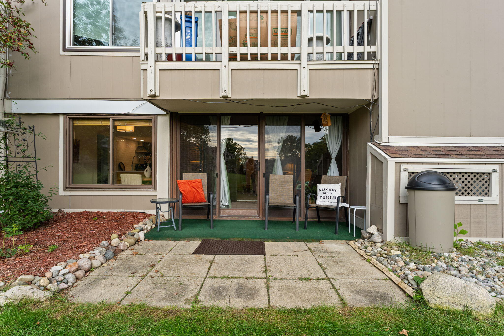 Condo patio with sliding doors, outdoor seating, and a green rug beside a stone-bordered walkway.