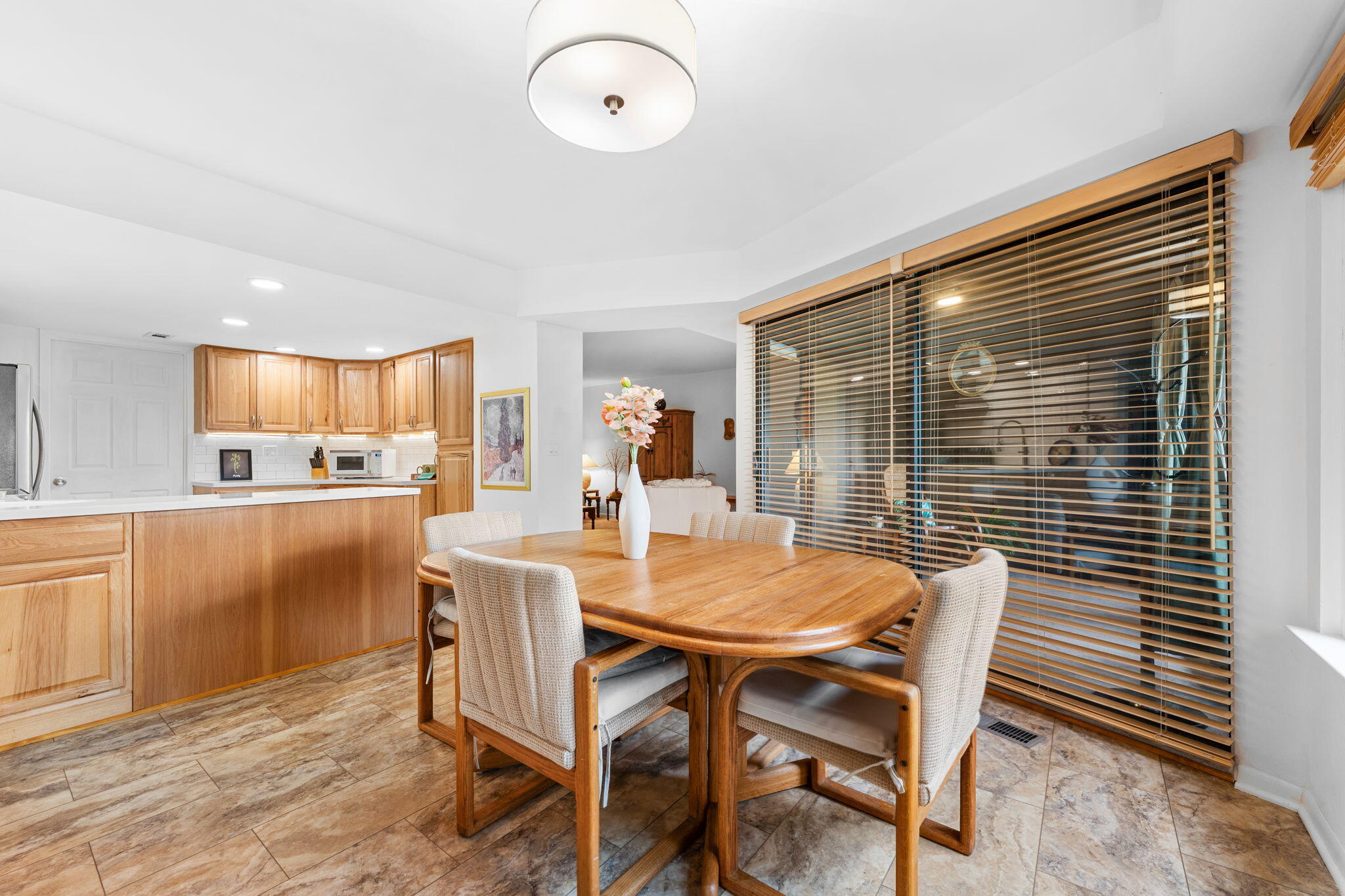 Dining area with an oval wooden table beside a kitchen with wood cabinets and large windows.