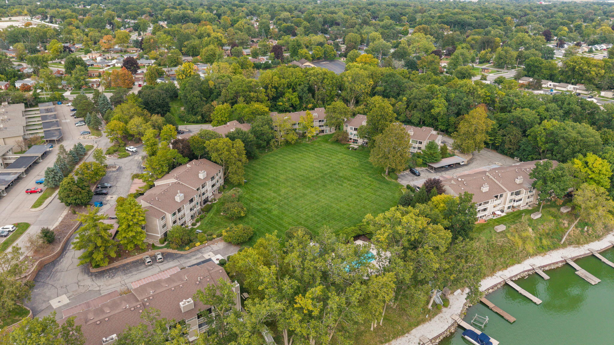 Aerial view of lakeside condominiums surrounding a large lawn, with mature trees and docks along the water.