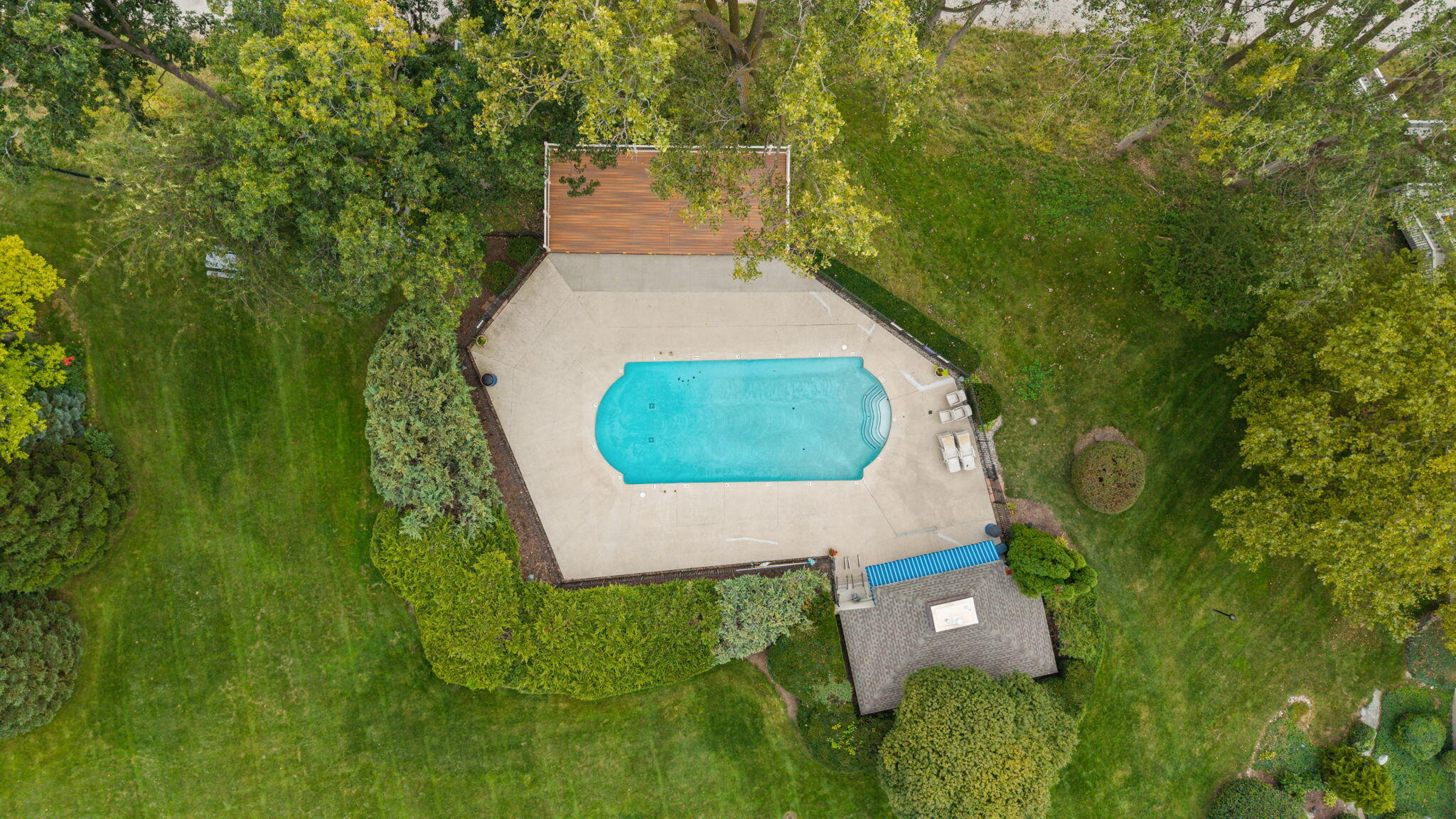 Outdoor swimming pool with a broad concrete deck, lounge chairs, and surrounding mature trees.