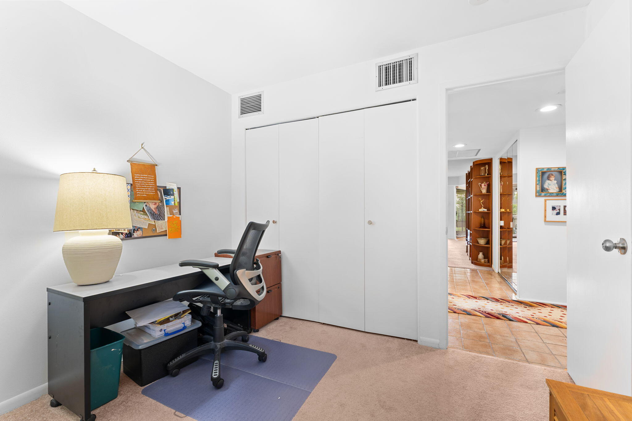 Home office with a black desk and ergonomic chair beside white closet doors and an open tiled hallway.
